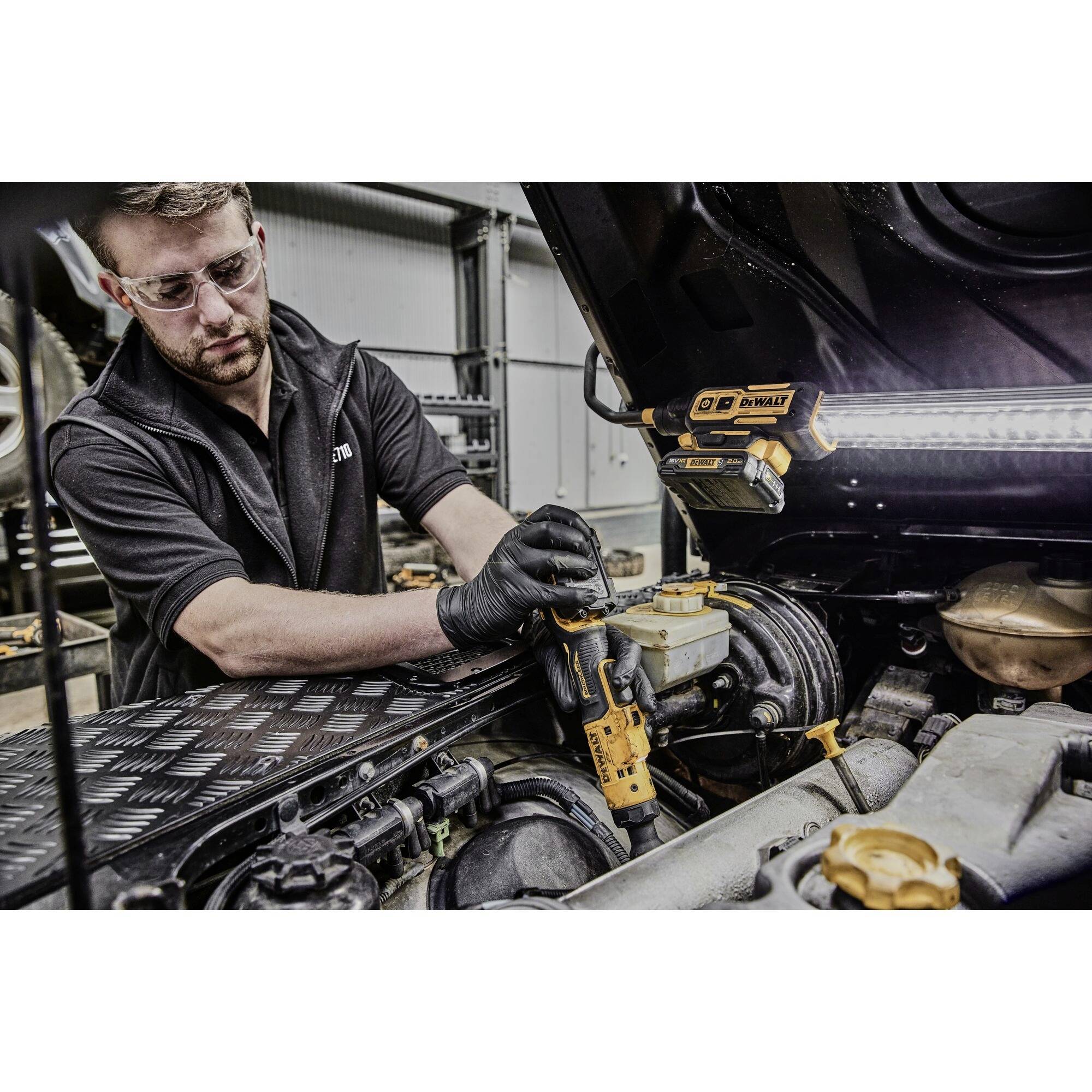 A man in workshop attire is repairing the interior of an open car boot with a screwdriver. Safety glasses and lighting are present.