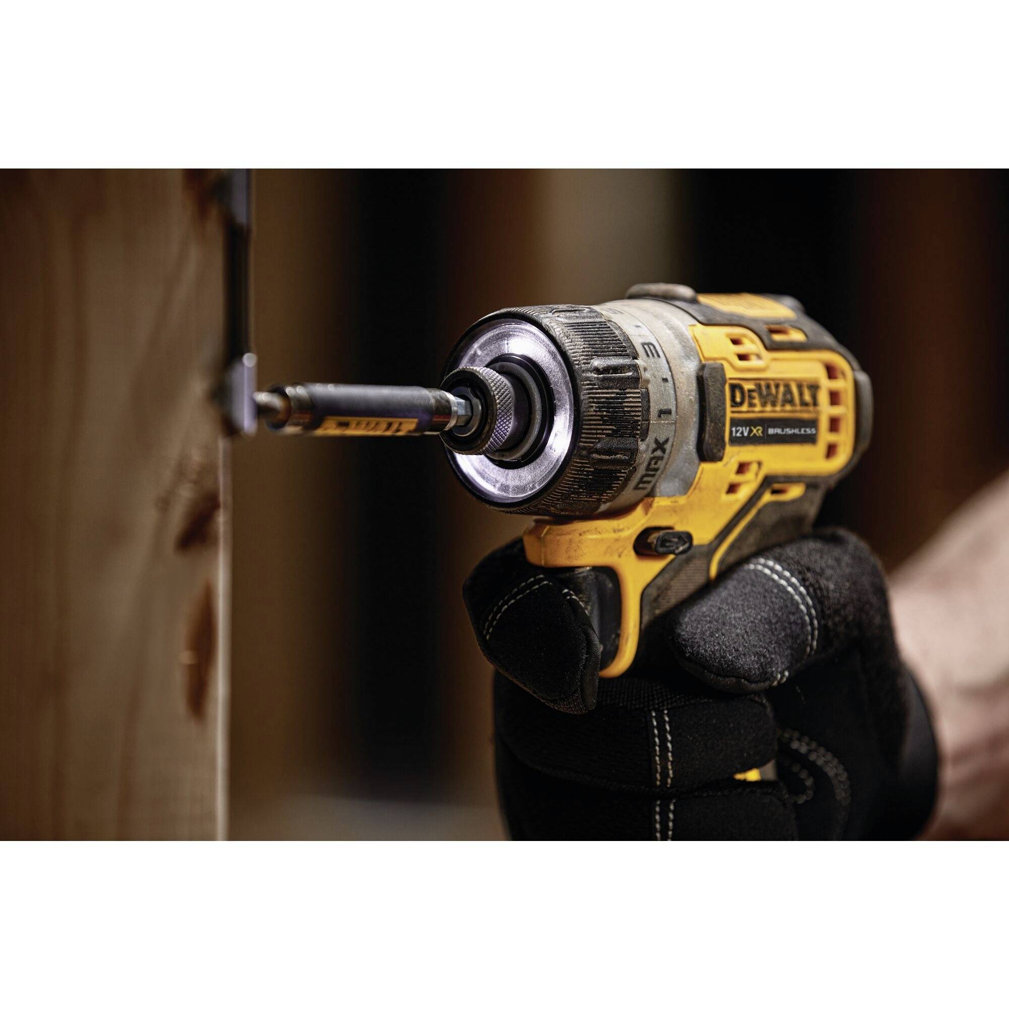 A person is drilling into a wooden board with a yellow cordless drill. The drill is shown in close-up, with the hand wearing a black glove.