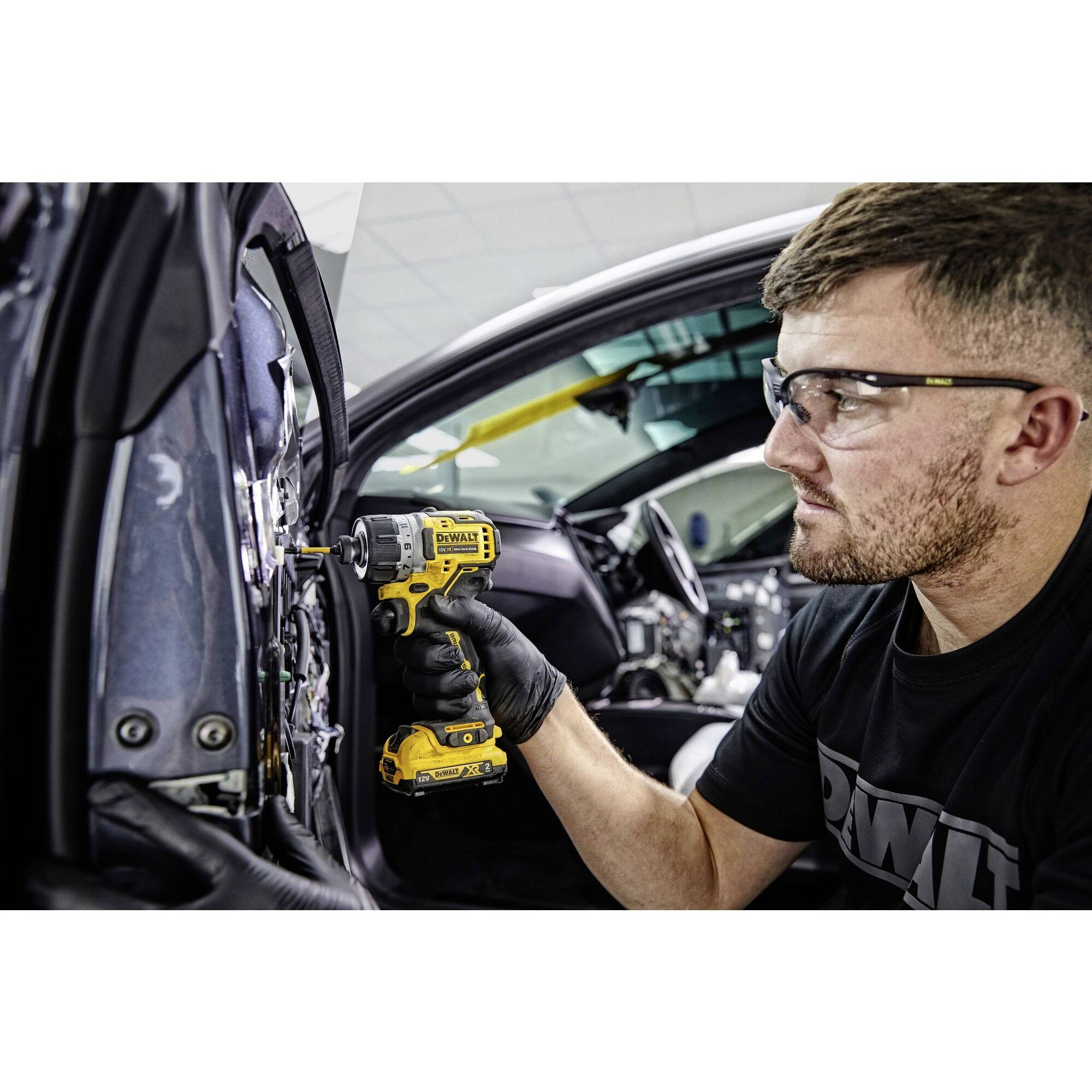 A man wearing safety glasses is using a yellow cordless drill to work on a car. The interior of the car is visible in the background.