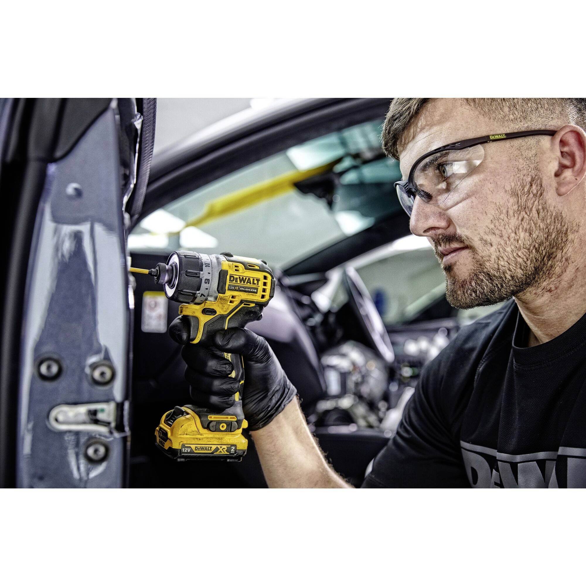 A person is repairing a car with an electric screwdriver. They are wearing safety glasses and gloves. The car is open.