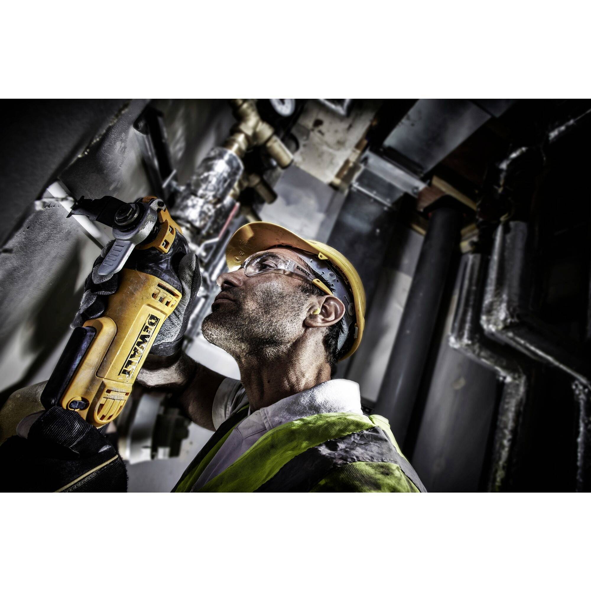 A worker wearing a hard hat and safety glasses uses an electric tool on a wall. He is wearing a high-visibility vest. Working in a technical environment.
