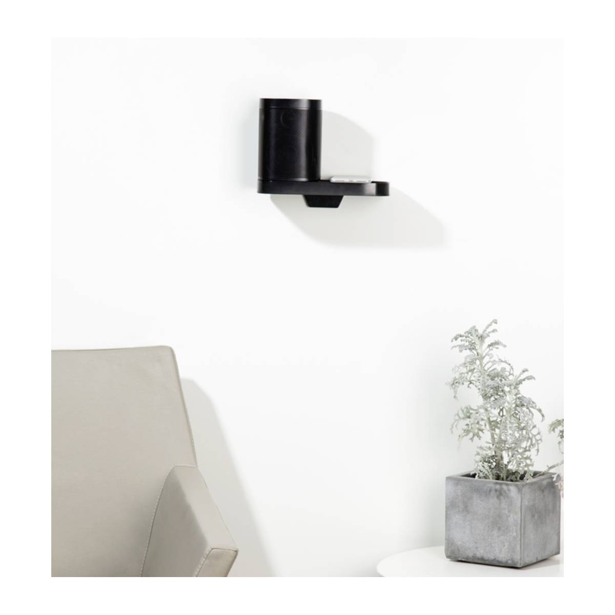 Black wall shelf with a round vase on top. Next to it, a corner sofa and a small, white table with a potted plant. Light-coloured wall in the background.