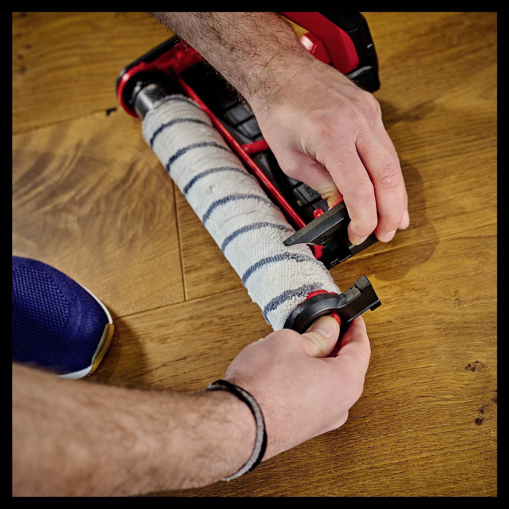 A person is changing the roller brush of a vacuum cleaner on a wooden floor, holding the end caps of the brush roller.