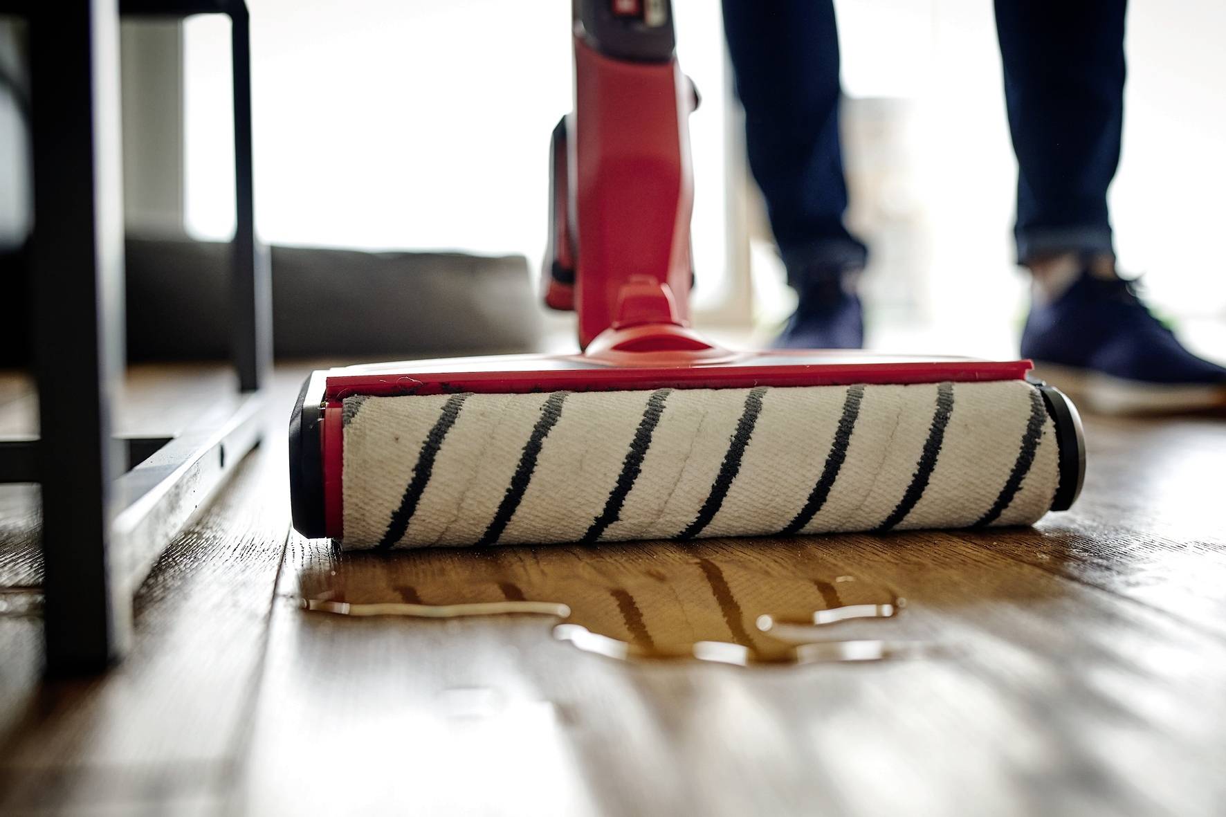 A mop is cleaning a wooden floor, with water spread across the surface. Blurred legs of a person can be seen in the background.