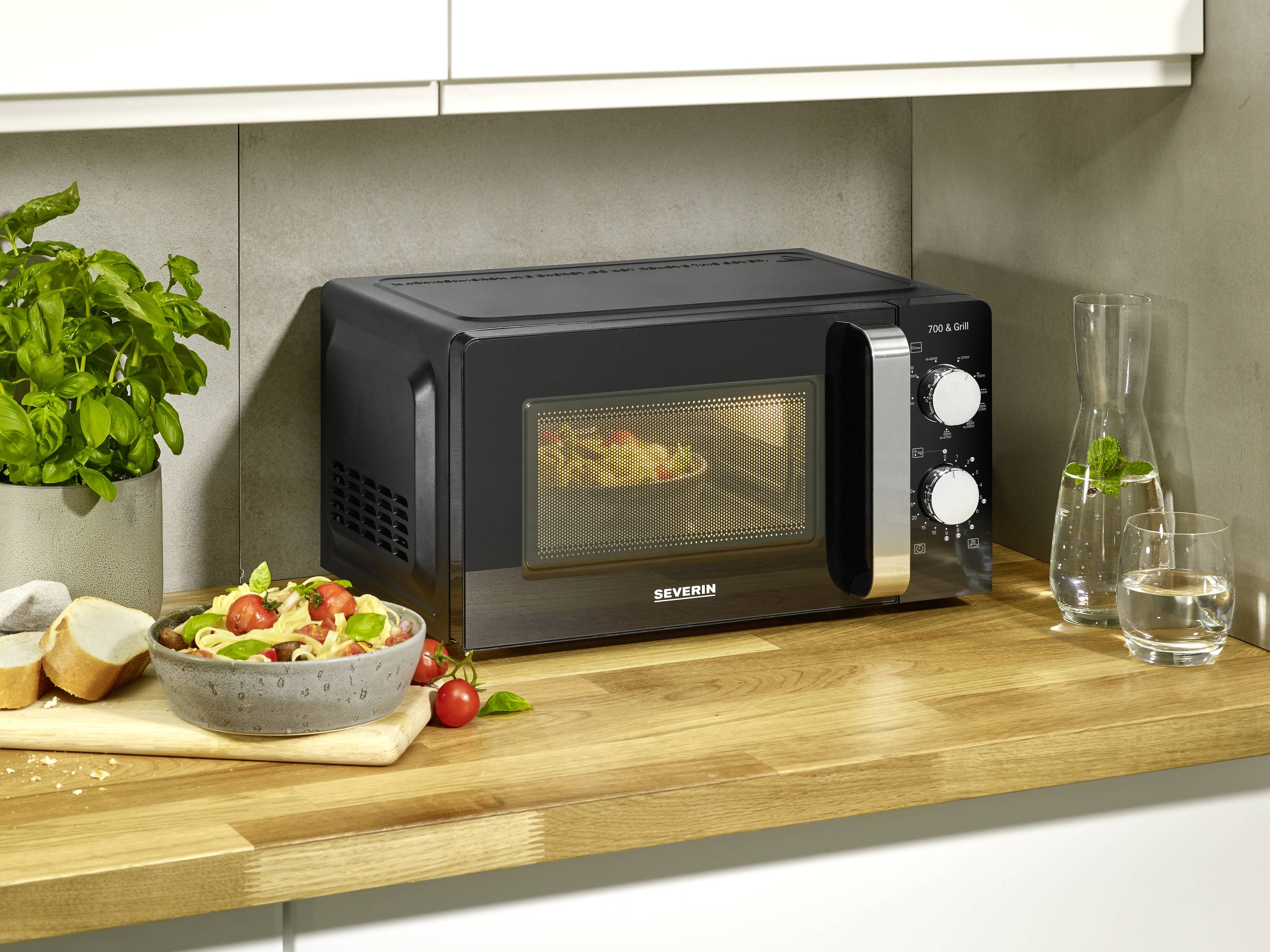 A black microwave oven sits on a kitchen worktop next to a salad and bread. In the background, a plant and water jugs are visible.