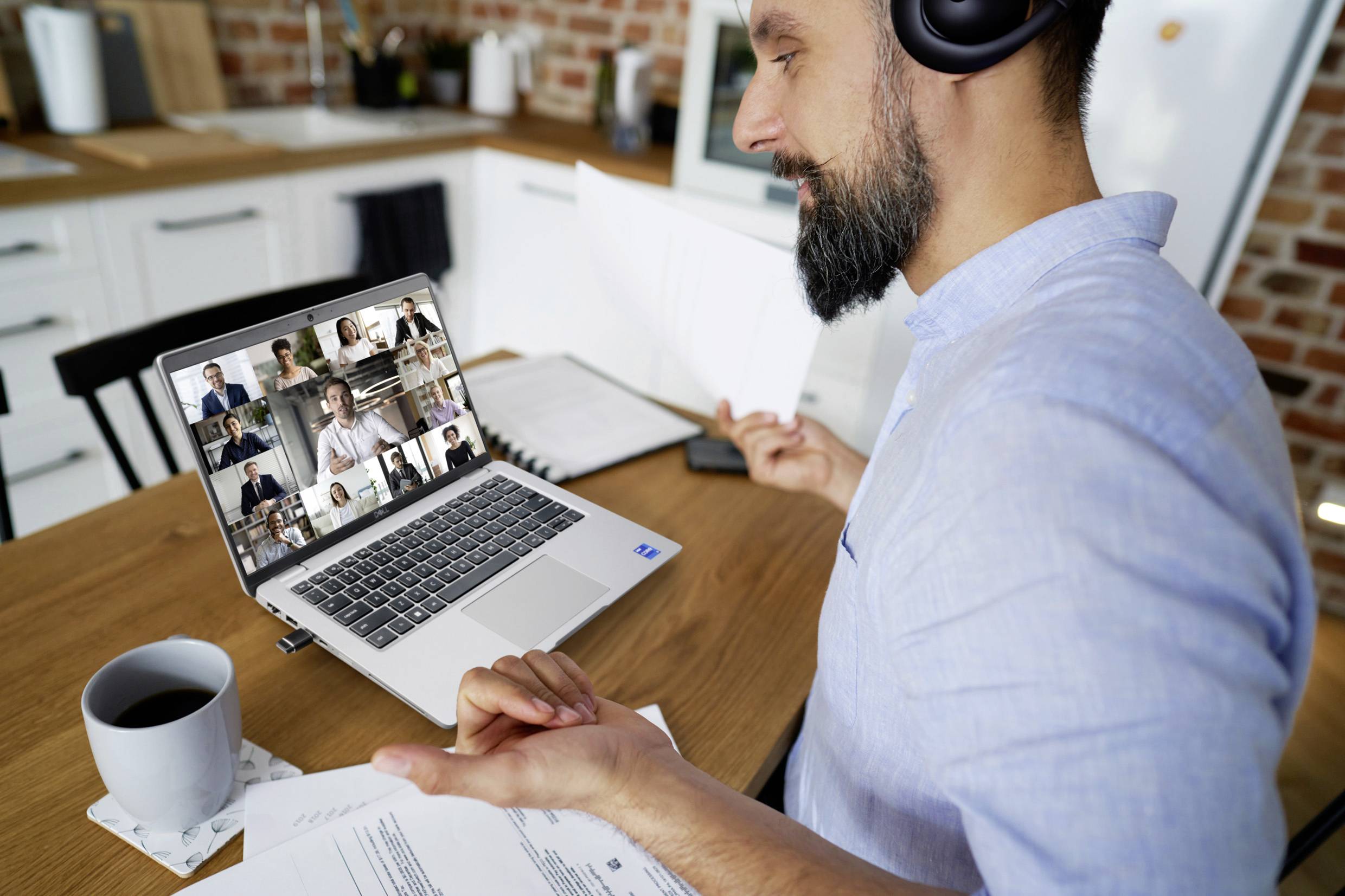 A man wearing headphones sits at a table, holding documents while participating in a video conference with multiple people on a laptop.