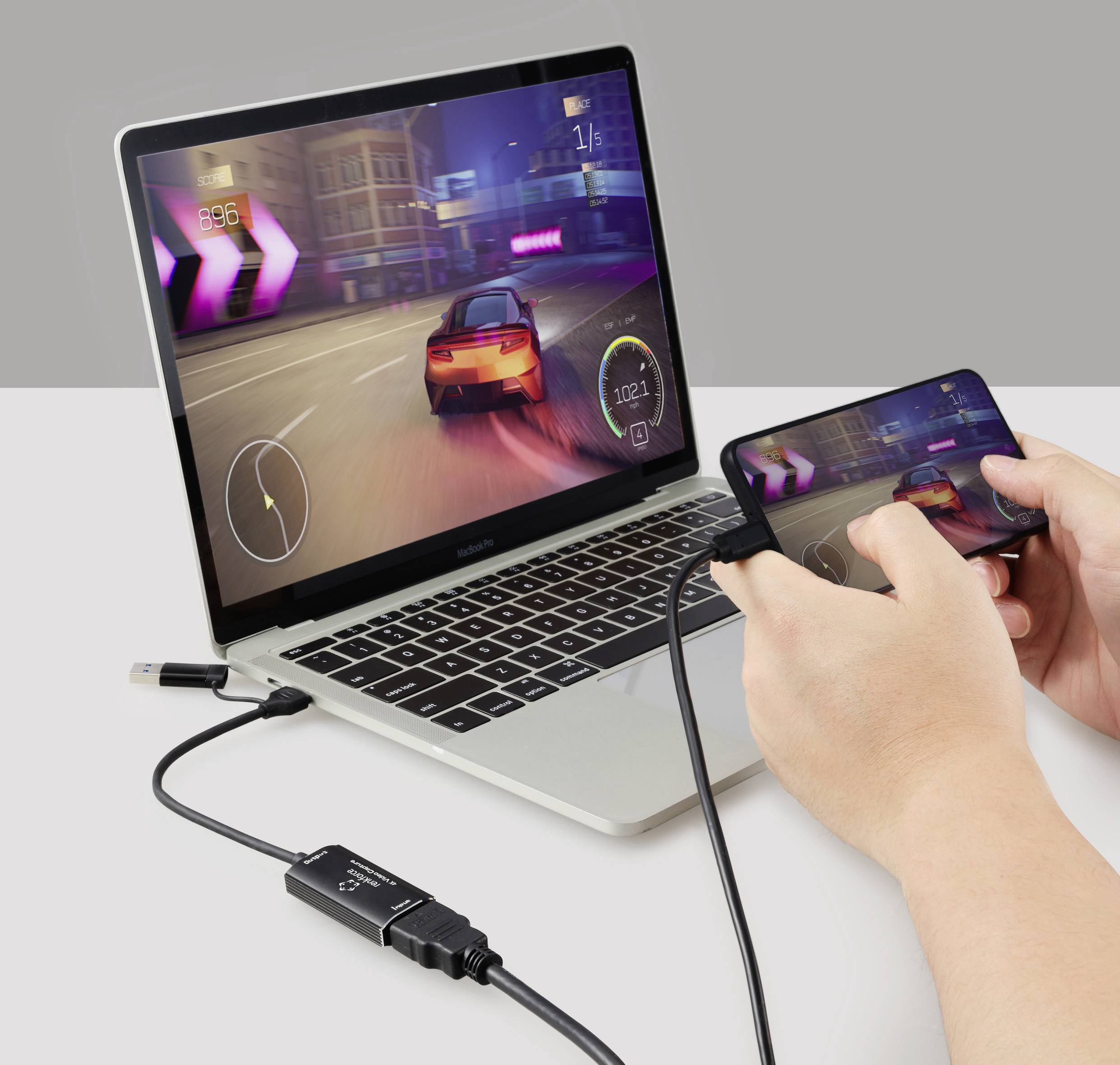 A person is playing a racing game on a smartphone connected to a laptop via cable. The laptop displays the same game.