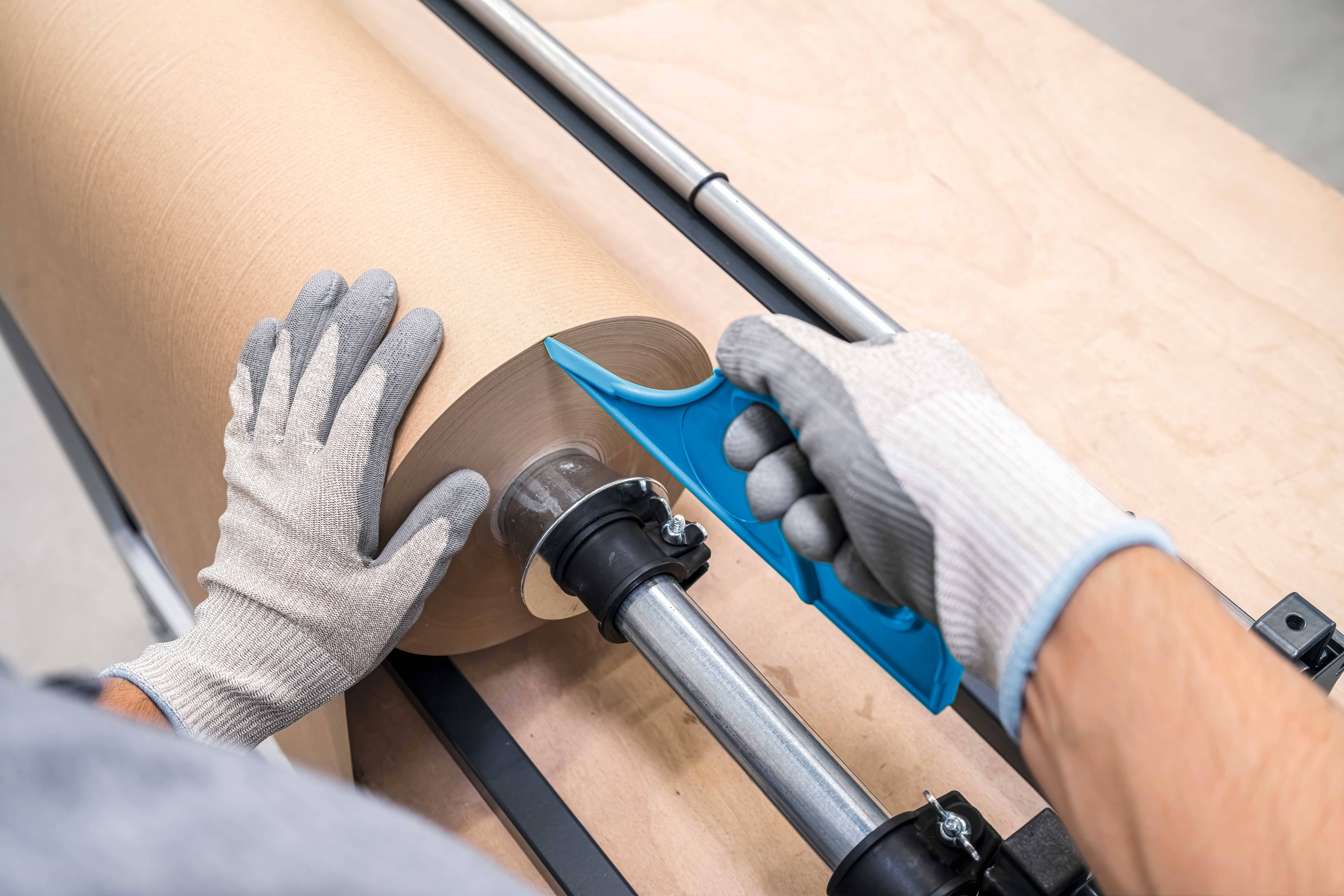 A person is cutting a large roll of brown paper with a blue tool on a cutting table. They are wearing grey gloves.
