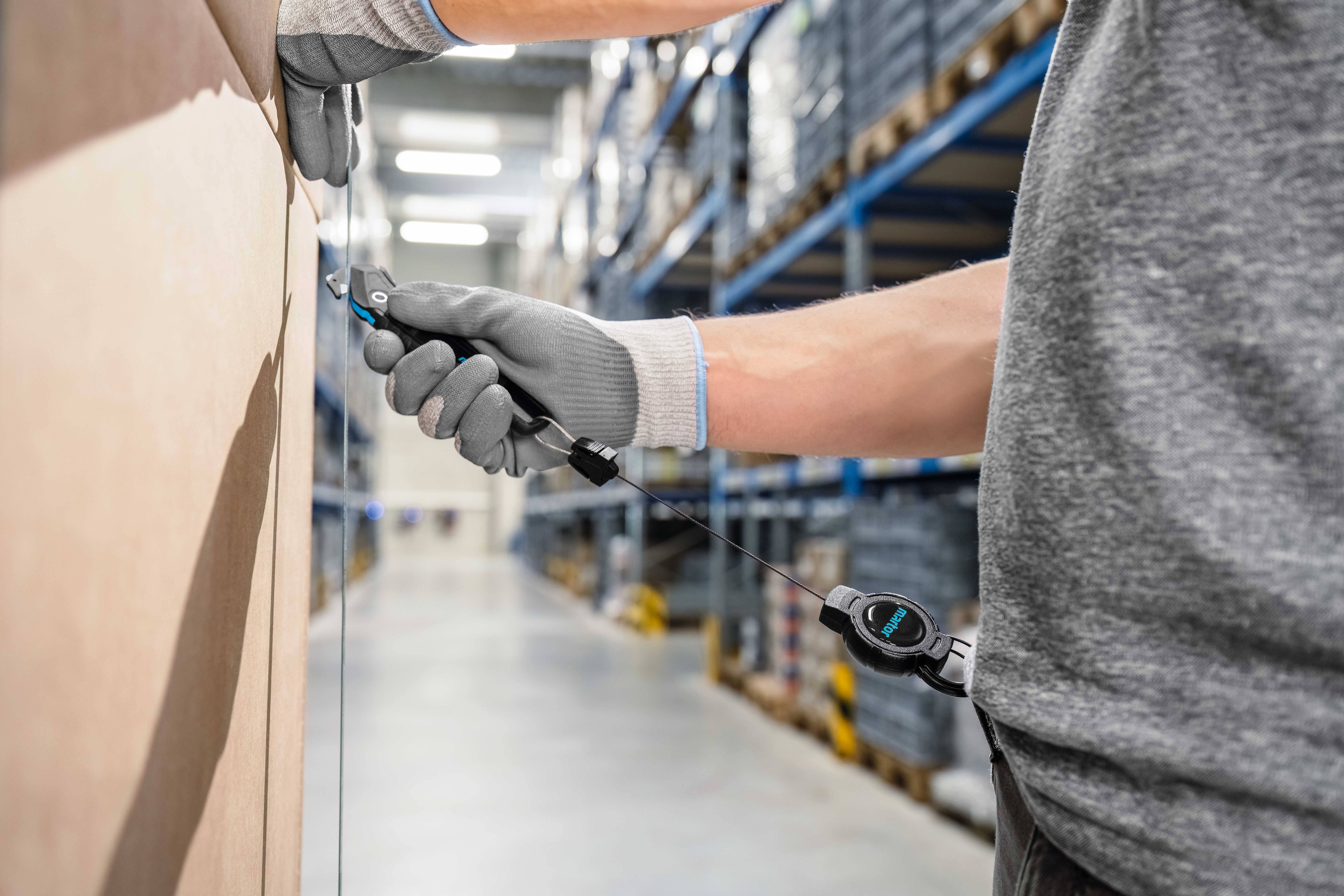 A person wearing a grey shirt and gloves is cutting cardboard with a utility knife in a warehouse. Storage shelves are visible in the background.