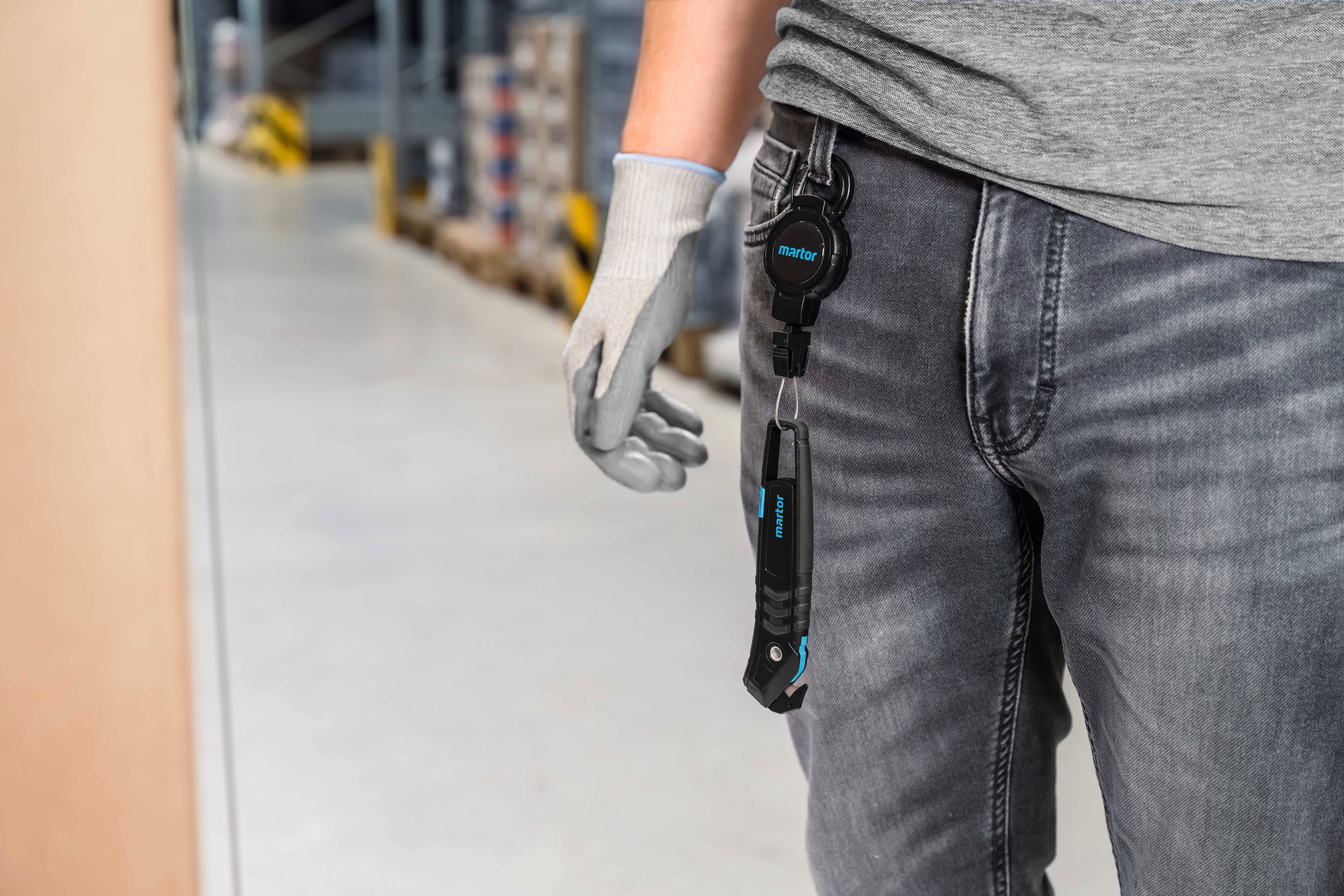 A person in the warehouse is wearing a belt with attached tools. They are wearing grey gloves and jeans. The background shows shelves and boxes.