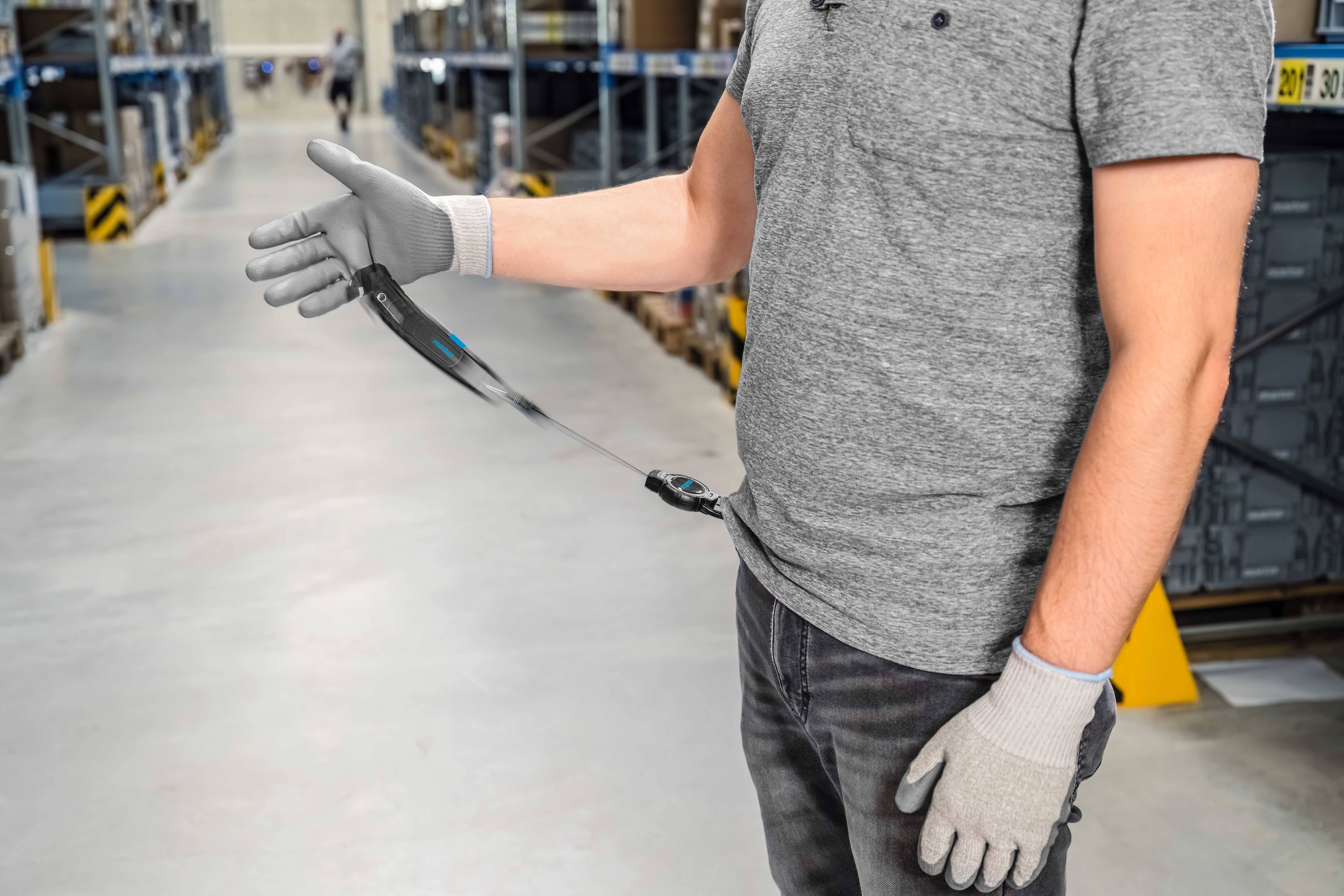 A person wearing a grey T-shirt and grey gloves is standing in a warehouse, with a safety line attached to their belt.