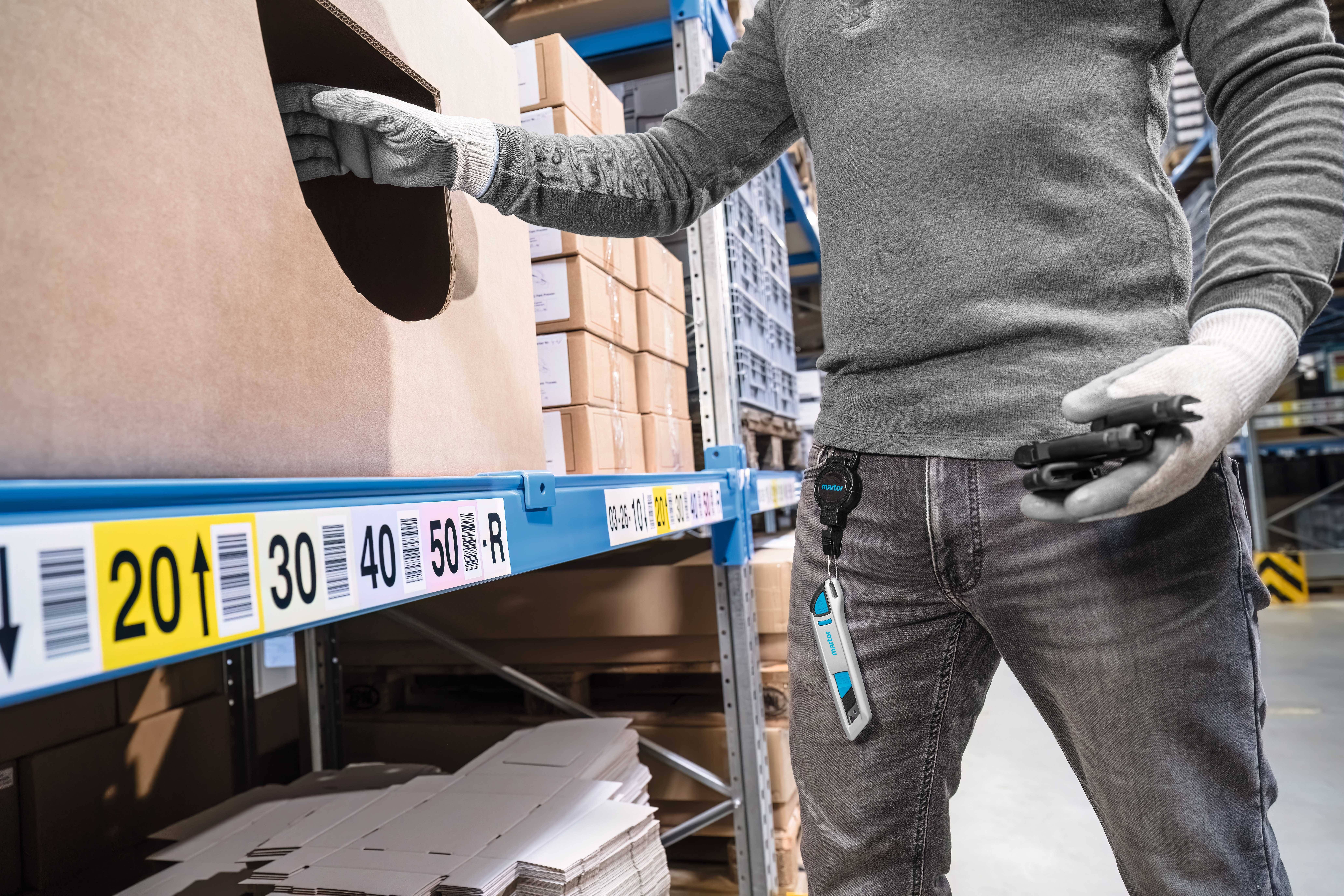 A person wearing gloves is retrieving an item from a shelf in a warehouse. Shelves with cardboard boxes and labels with numbers are visible.