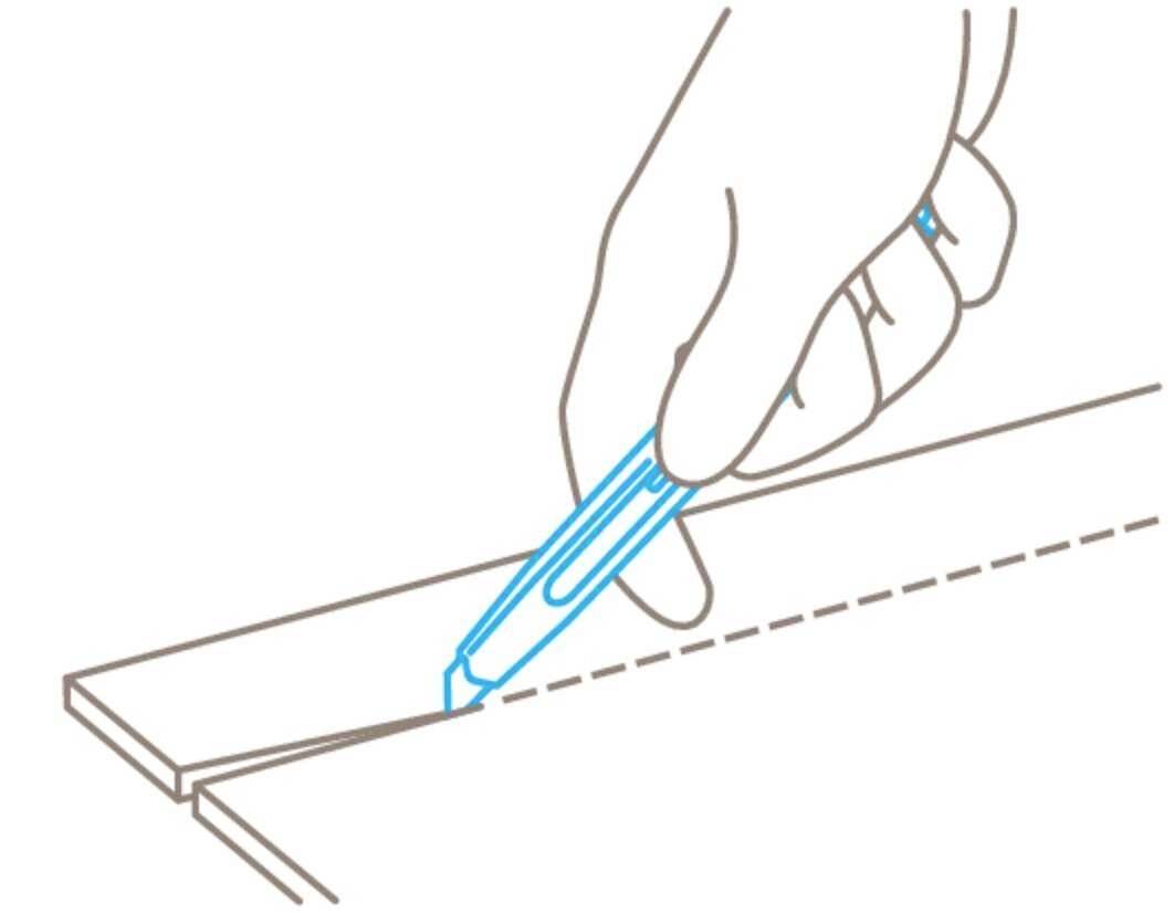 A hand is using a blue craft knife to cut along a dotted line on wood or cardboard.