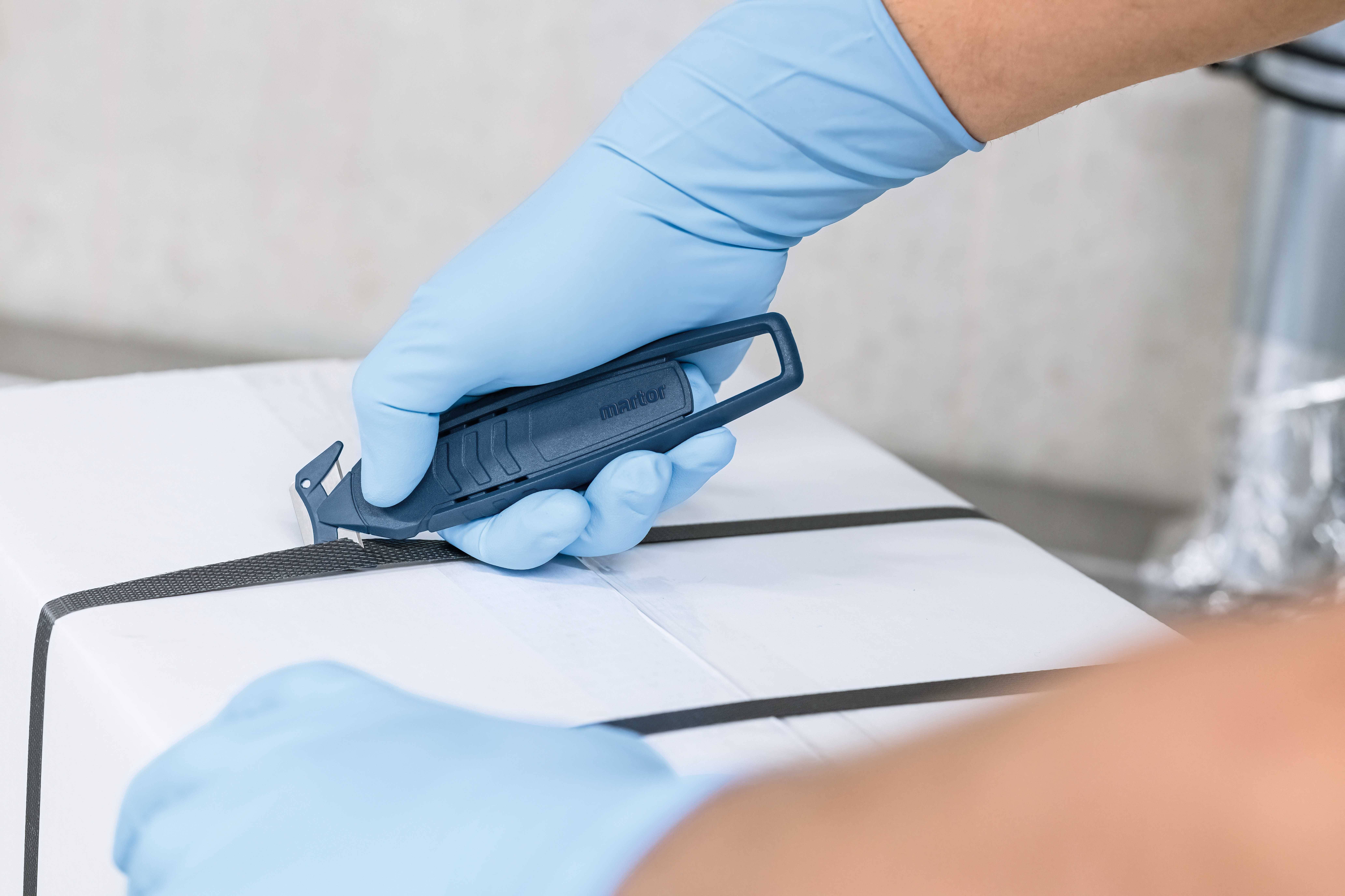 A person wearing blue gloves is cutting a black ribbon on a white package with a blue safety knife.