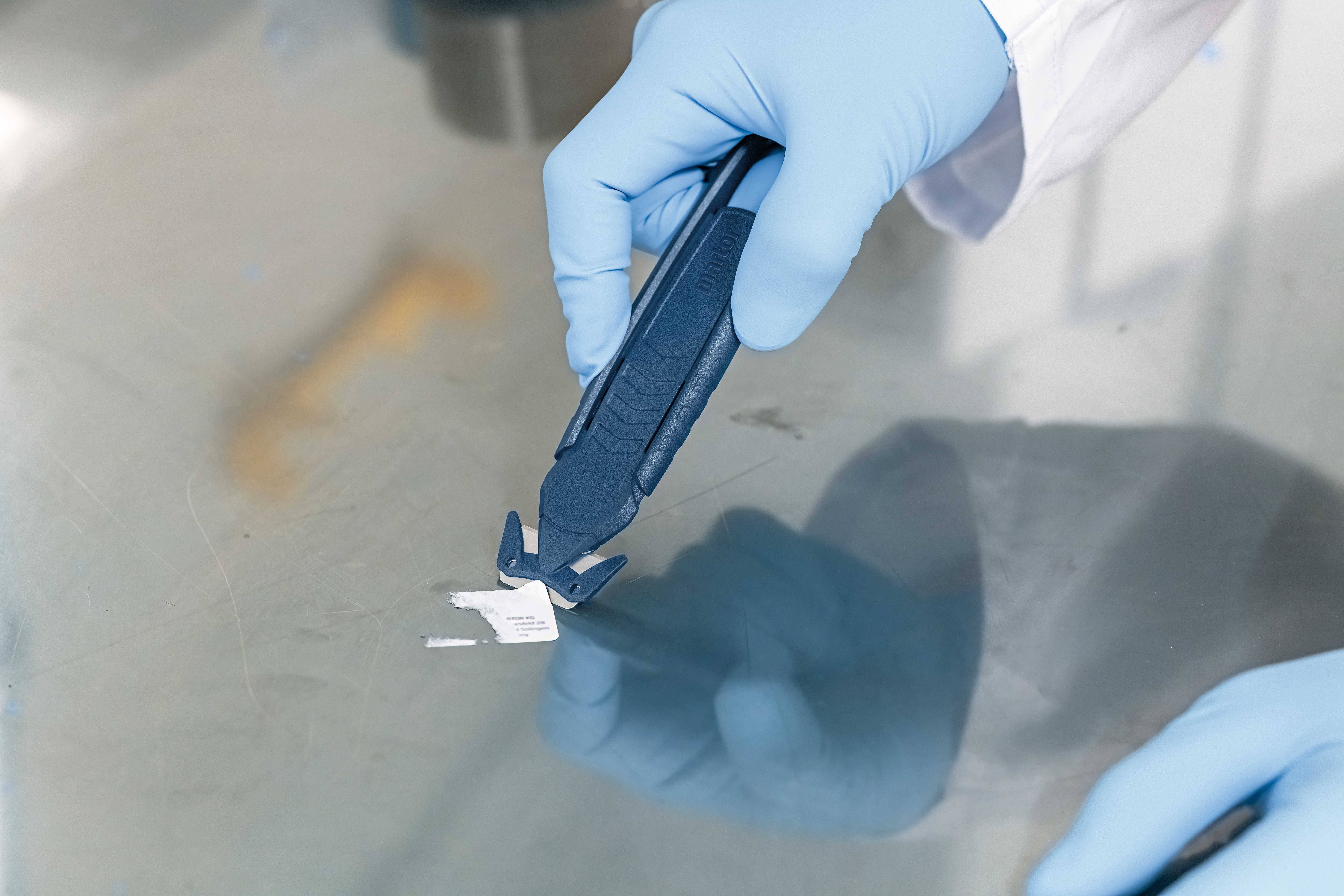 A hand wearing a blue glove holds a tool to remove a label from a metal surface.
