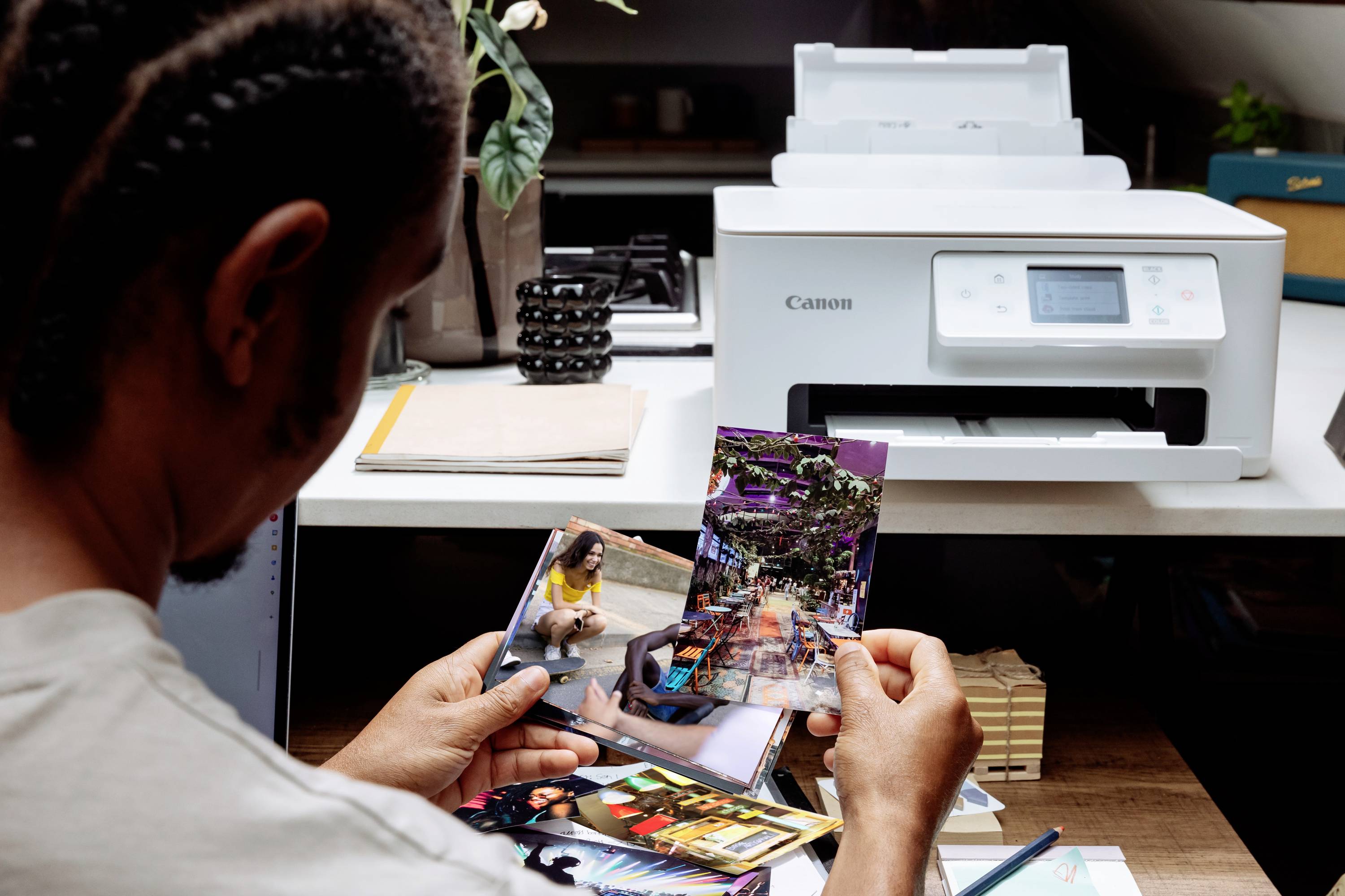 A person is examining photographs near a printer. The desk is covered with notebooks and a pen, with a focus on print quality.