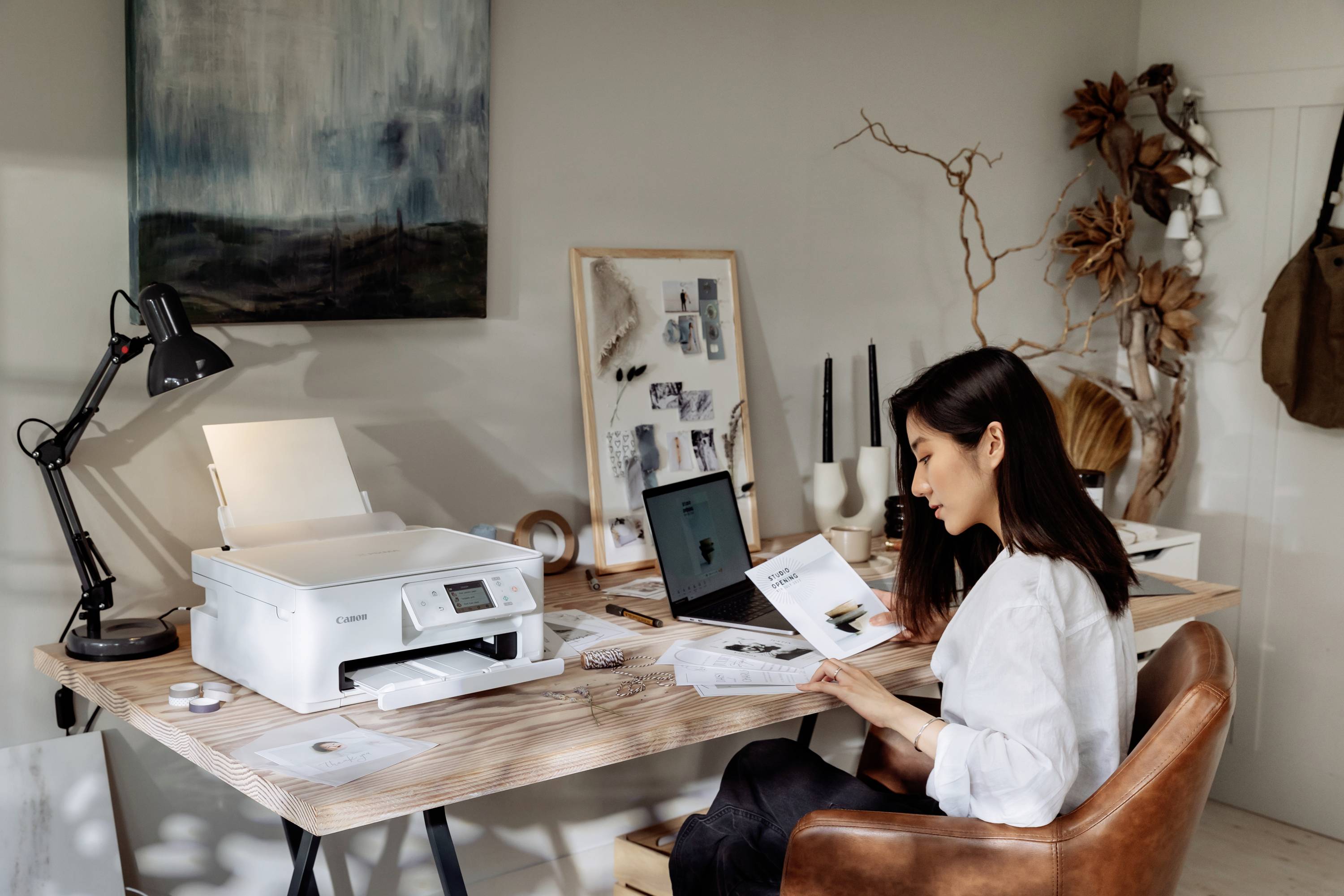 A woman is working at a wooden desk with a printer and laptop. She is reading a document attentively.