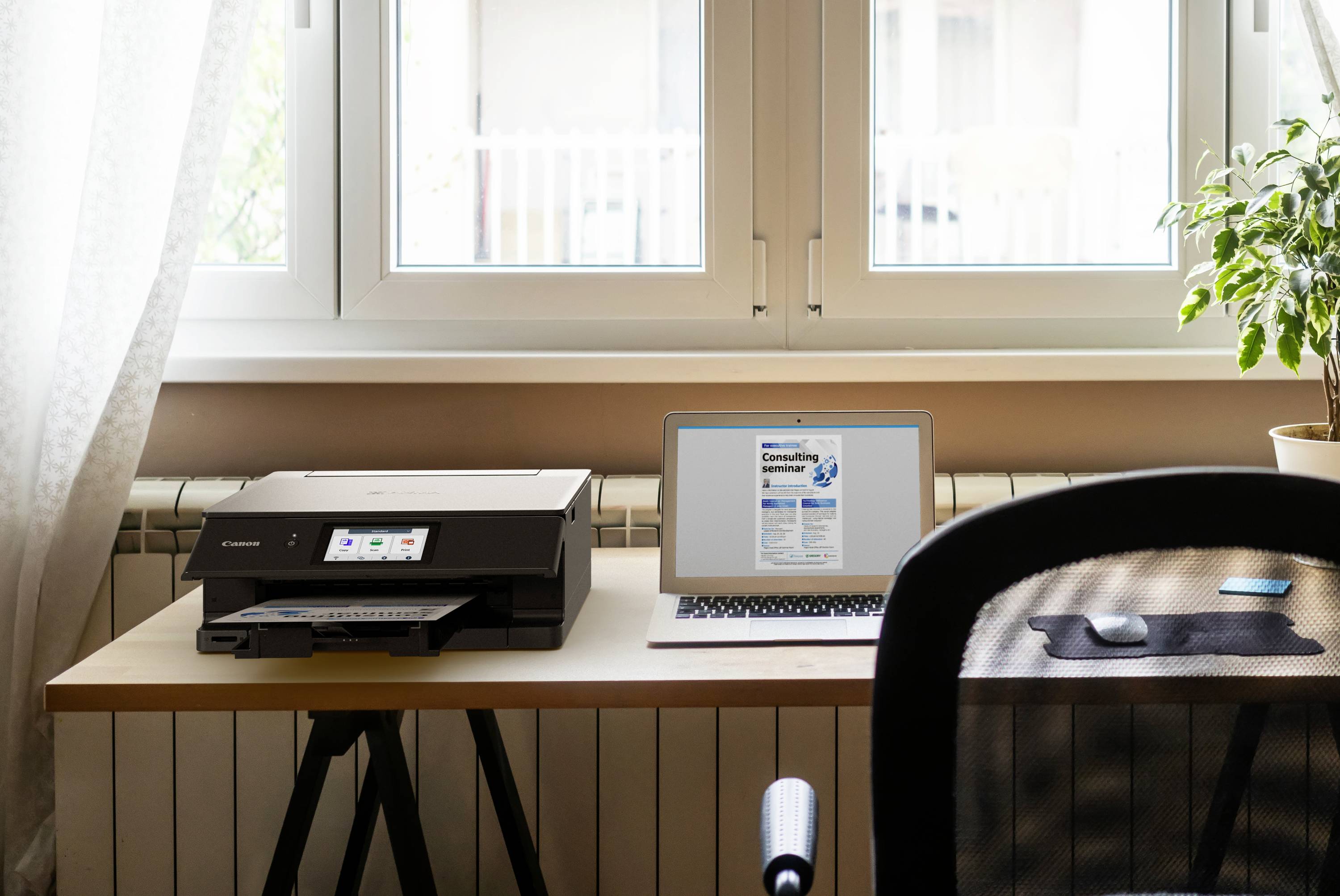 Workspace with a laptop, printer, and potted plant on a table in front of a window. The laptop displays an open webpage.
