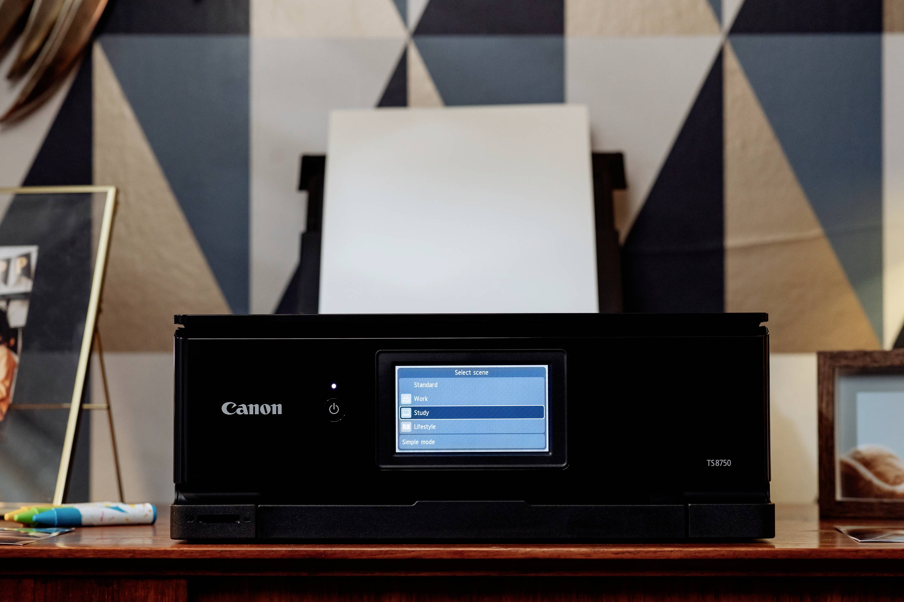 A printer on a table, surrounded by decorative frames. The screen displays a menu with options such as 'Photo' and 'Document'.