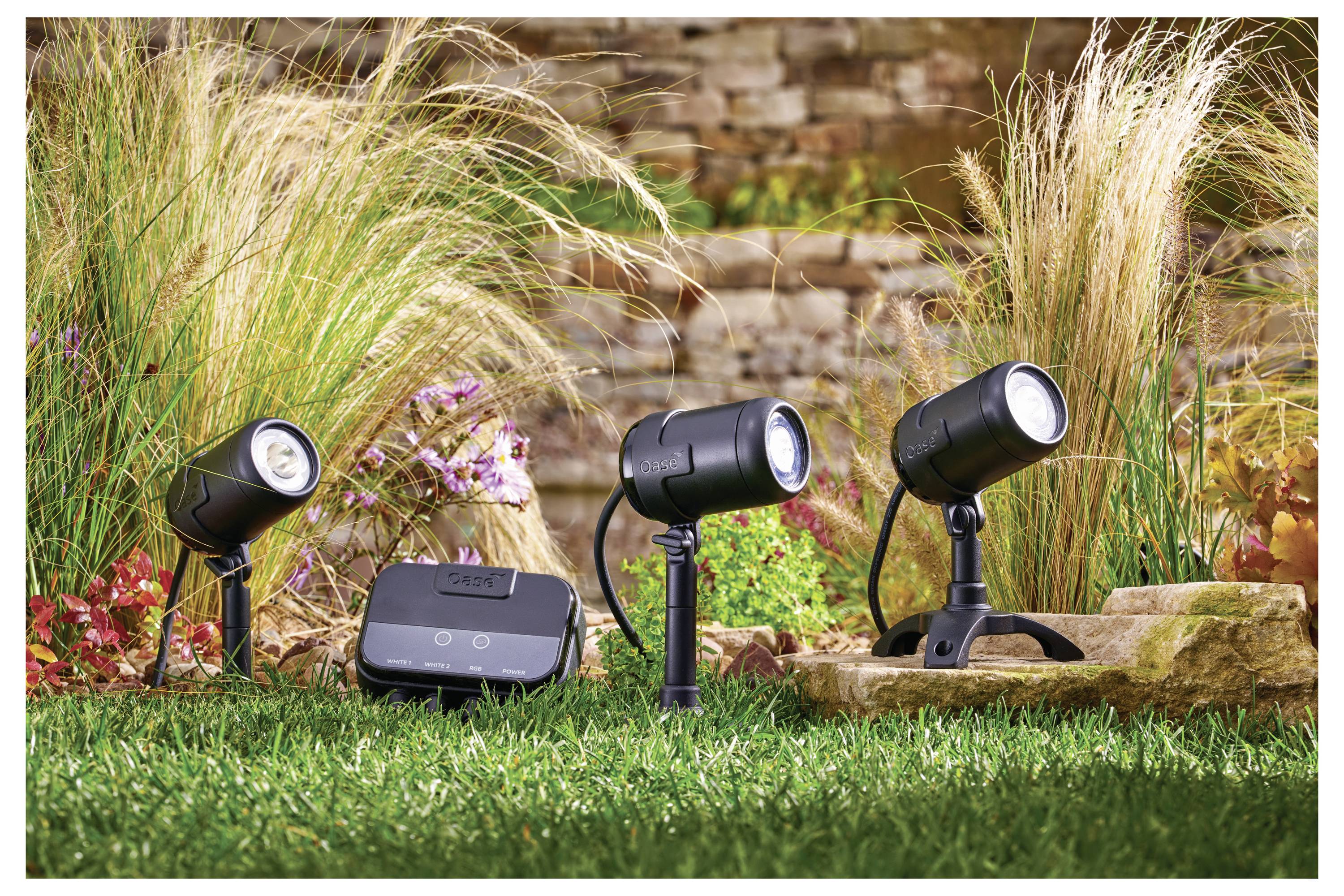 Outdoor landscape lighting setup on grass with three spotlights and a control unit, surrounded by ornamental grass and stone wall.
