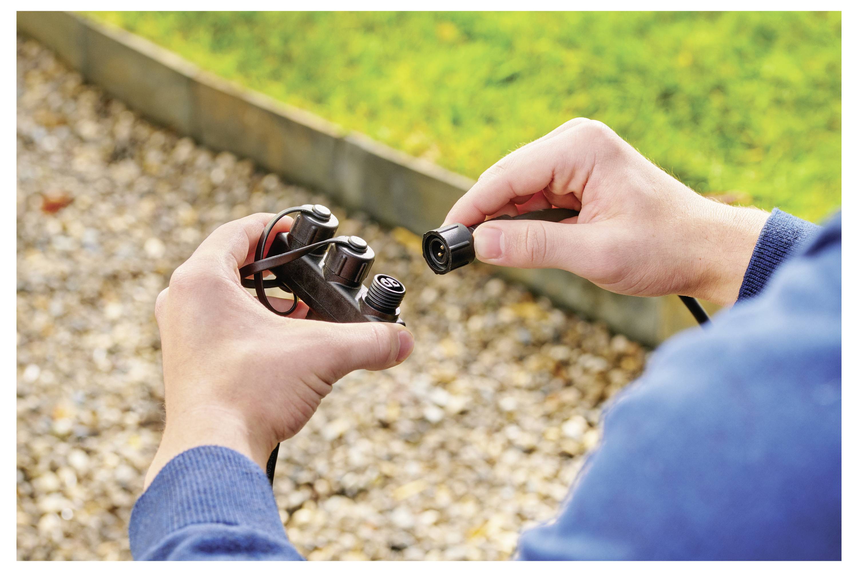 A person in a blue sweater connects a garden hose to a watering system control unit set over pebbled ground, with grass in the background.
