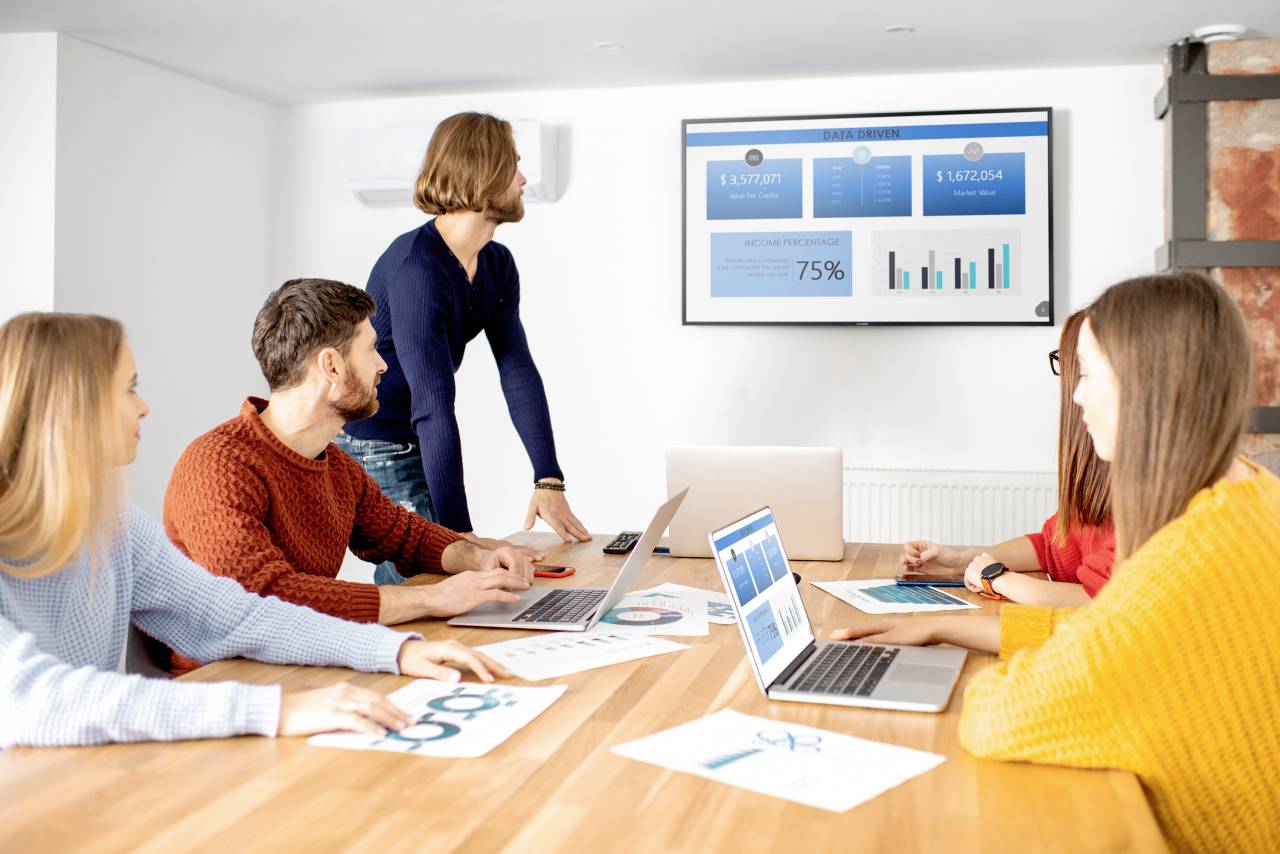 A group of five people are sitting around a table looking at a screen with financial charts and data in a meeting room.