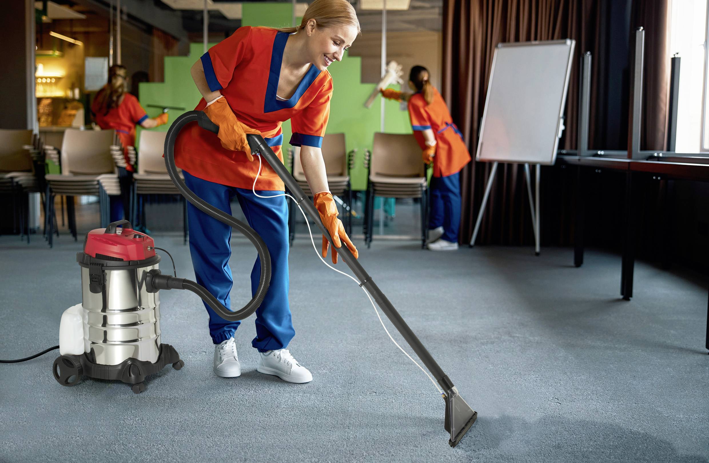 A woman in work attire is vacuuming the carpet with an industrial vacuum cleaner in a conference room. In the background, another person is cleaning the space.