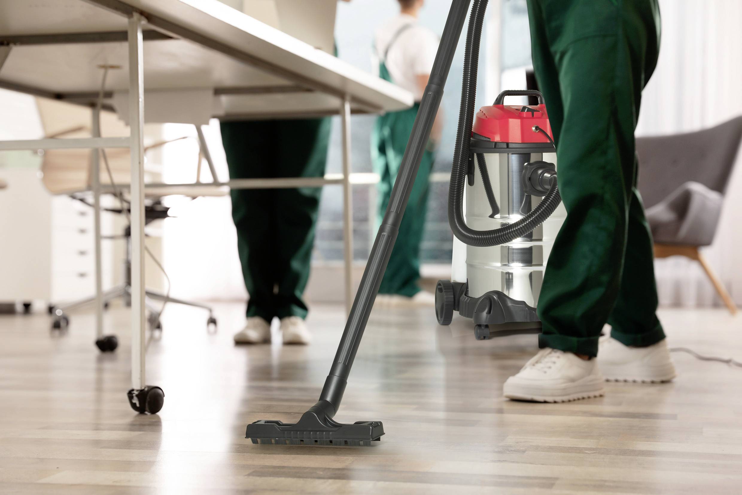 A person is cleaning an office floor using an industrial vacuum cleaner; other individuals can be seen performing office cleaning in the background.