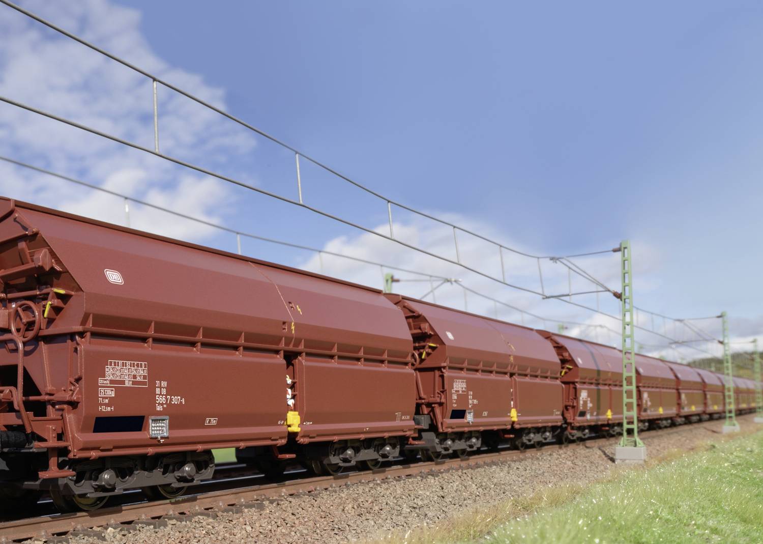 A long freight train with several red carriages travels along tracks beneath an electricity pylon through a rural landscape with a blue sky.