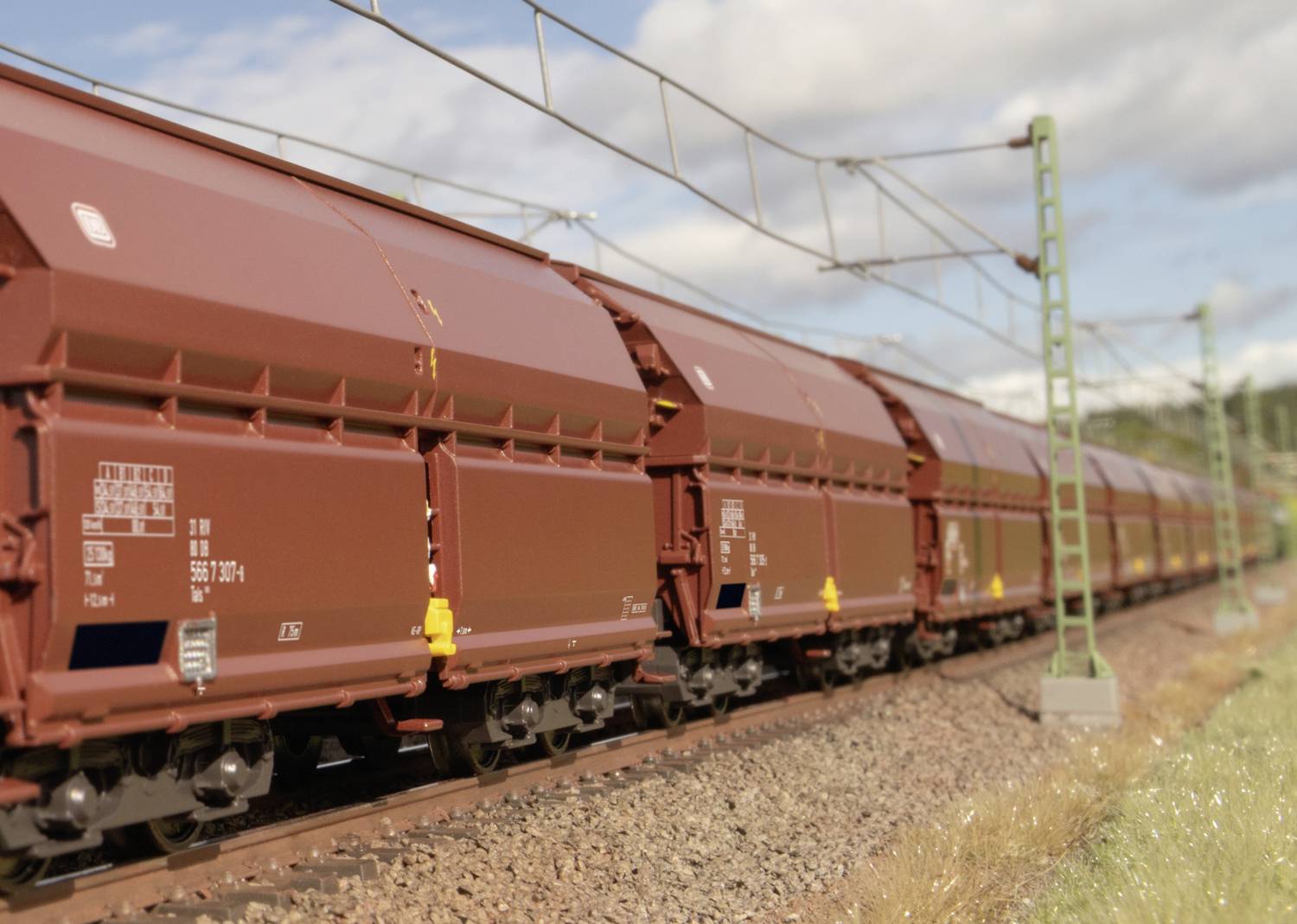 A long freight train with brown carriages travels along a track beneath an electricity transmission system in bright sunshine.
