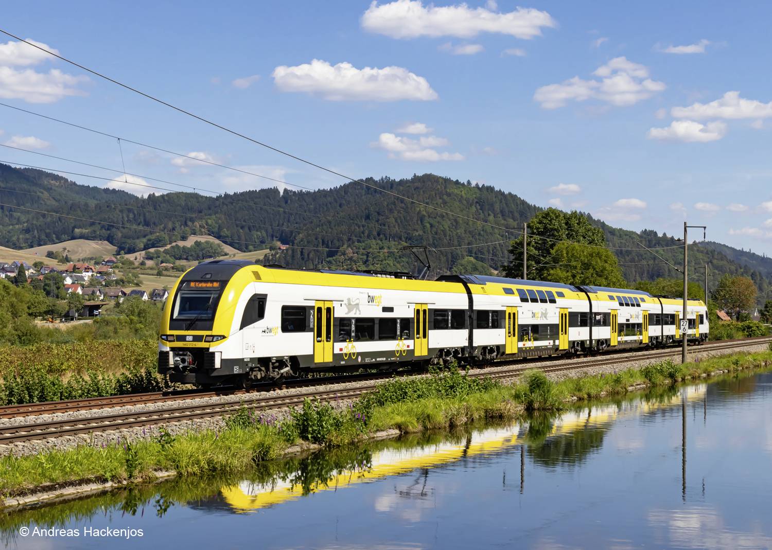 A yellow and white train travels through a rural landscape on a fine day, reflected in a water channel. Hills and clouds are visible in the background.