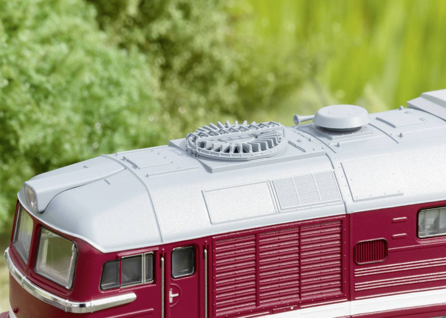 Red model of a locomotive in front of a blurred green background. Main details: Roof ventilation and window frames visible.