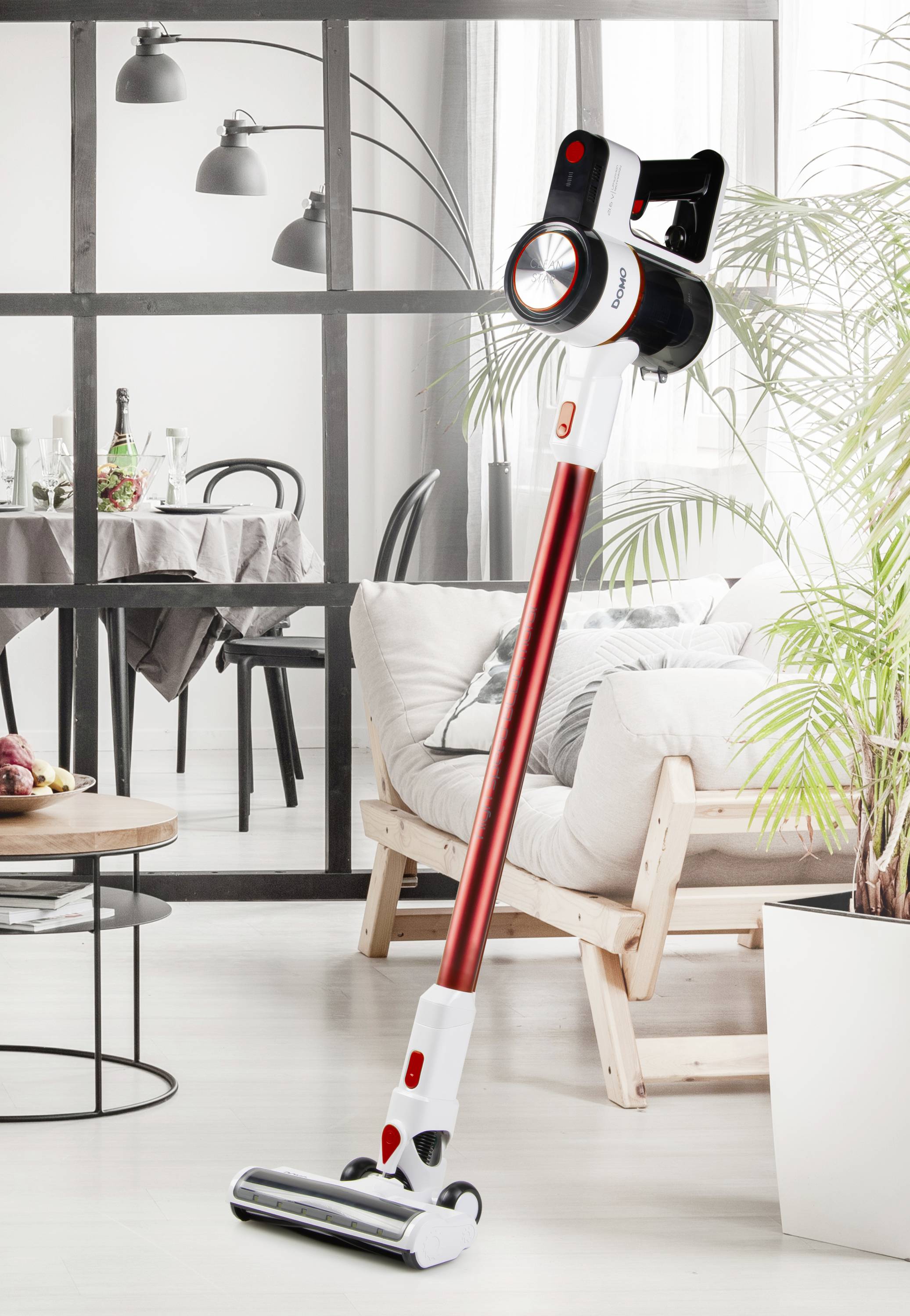 A cordless vacuum cleaner with a red handle stands upright in a modern living room with a light wooden chair, table, and plants.