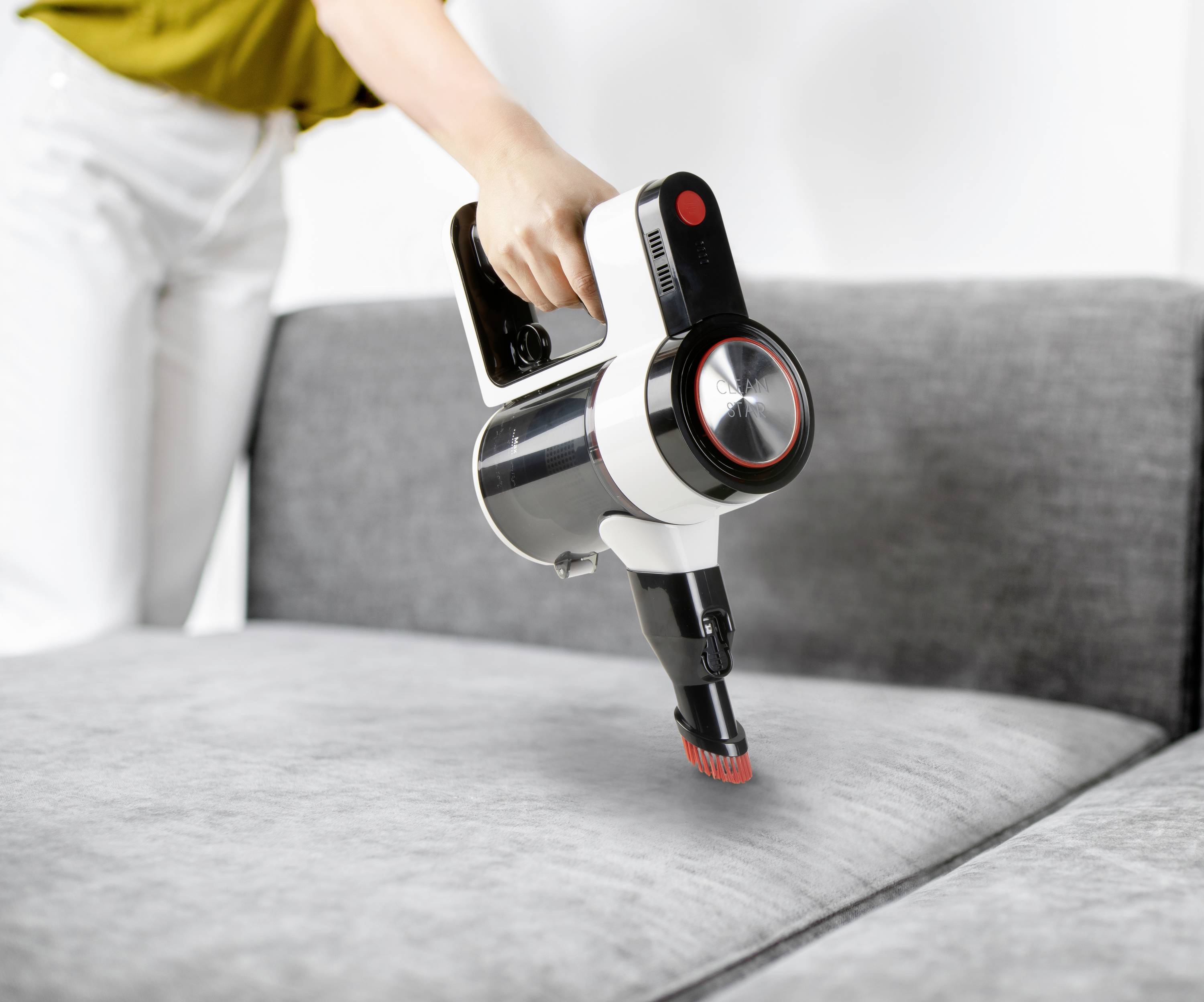 A person is cleaning a grey sofa with a cordless handheld vacuum cleaner.