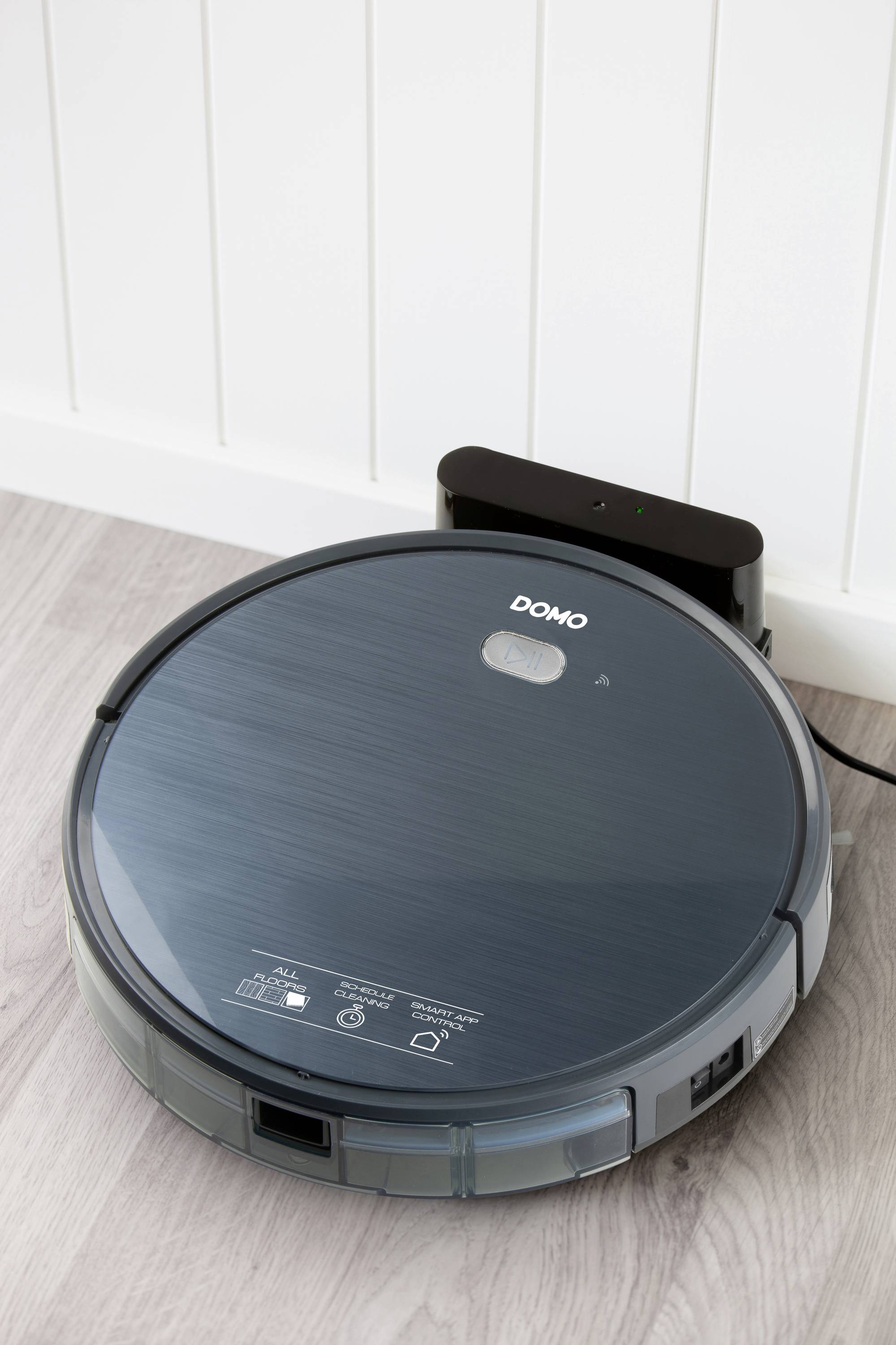 Robotic vacuum cleaner on a light wooden floor, docked at its charging station. Surface glossy, with brand logo visible. Background: white wall.