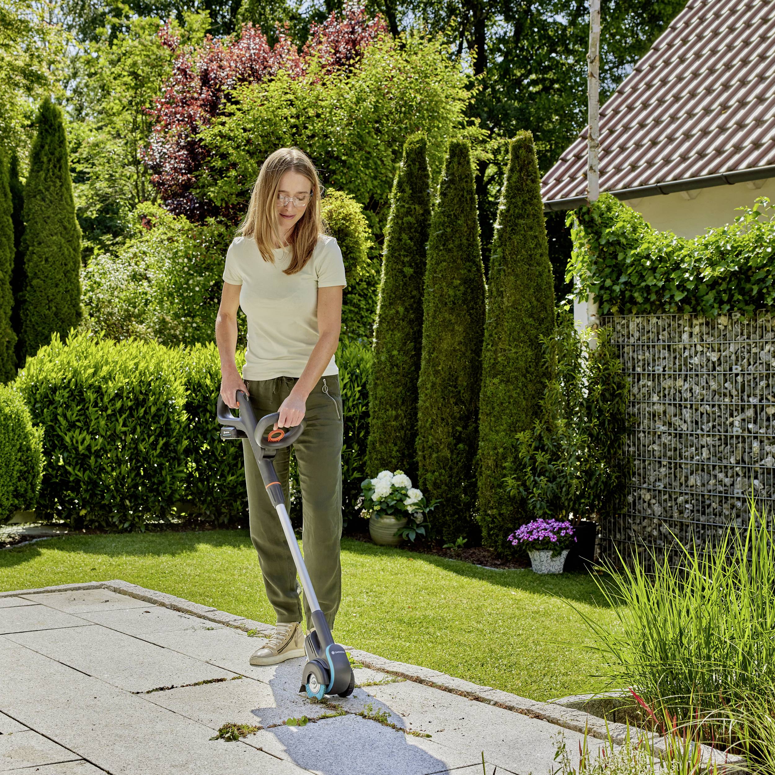 A woman is mowing the lawn in a sunny garden with an electric lawnmower. Trees and a house can be seen in the background.