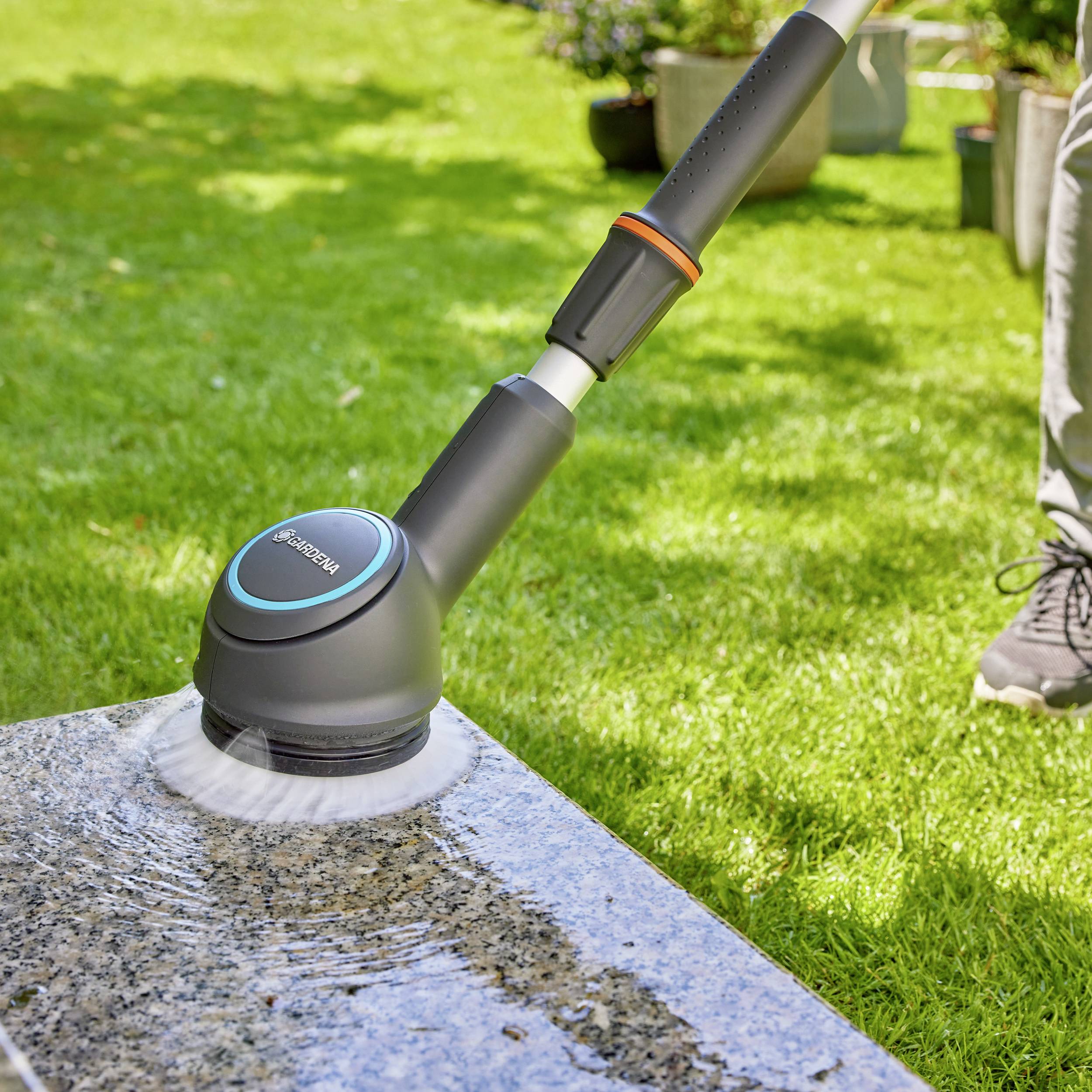 A cleaning brush with a water connection is cleaning a stone surface in the garden. In the background, grass and plants can be seen.