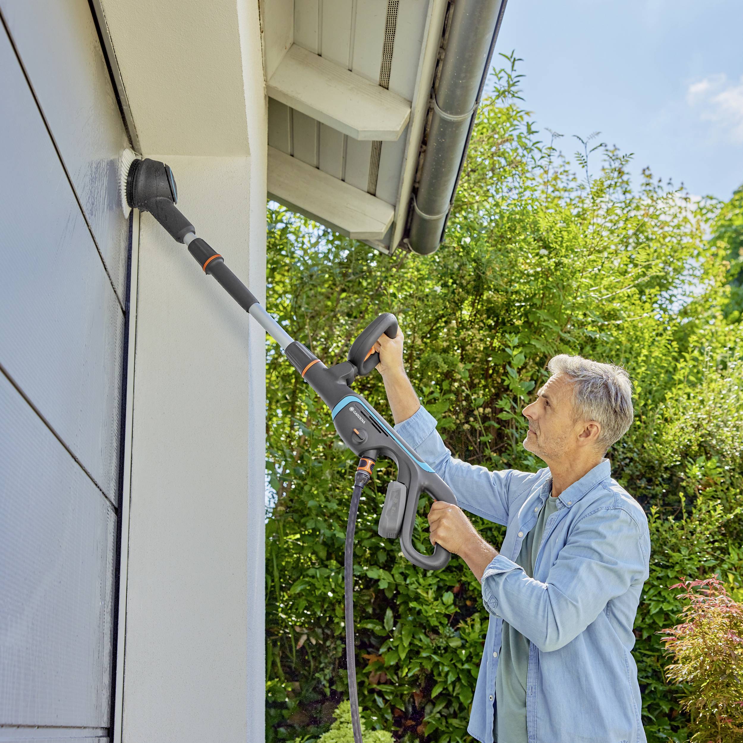 A person is cleaning the guttering of a house with a pressure washer in a green, rural setting. The sky is partly cloudy.