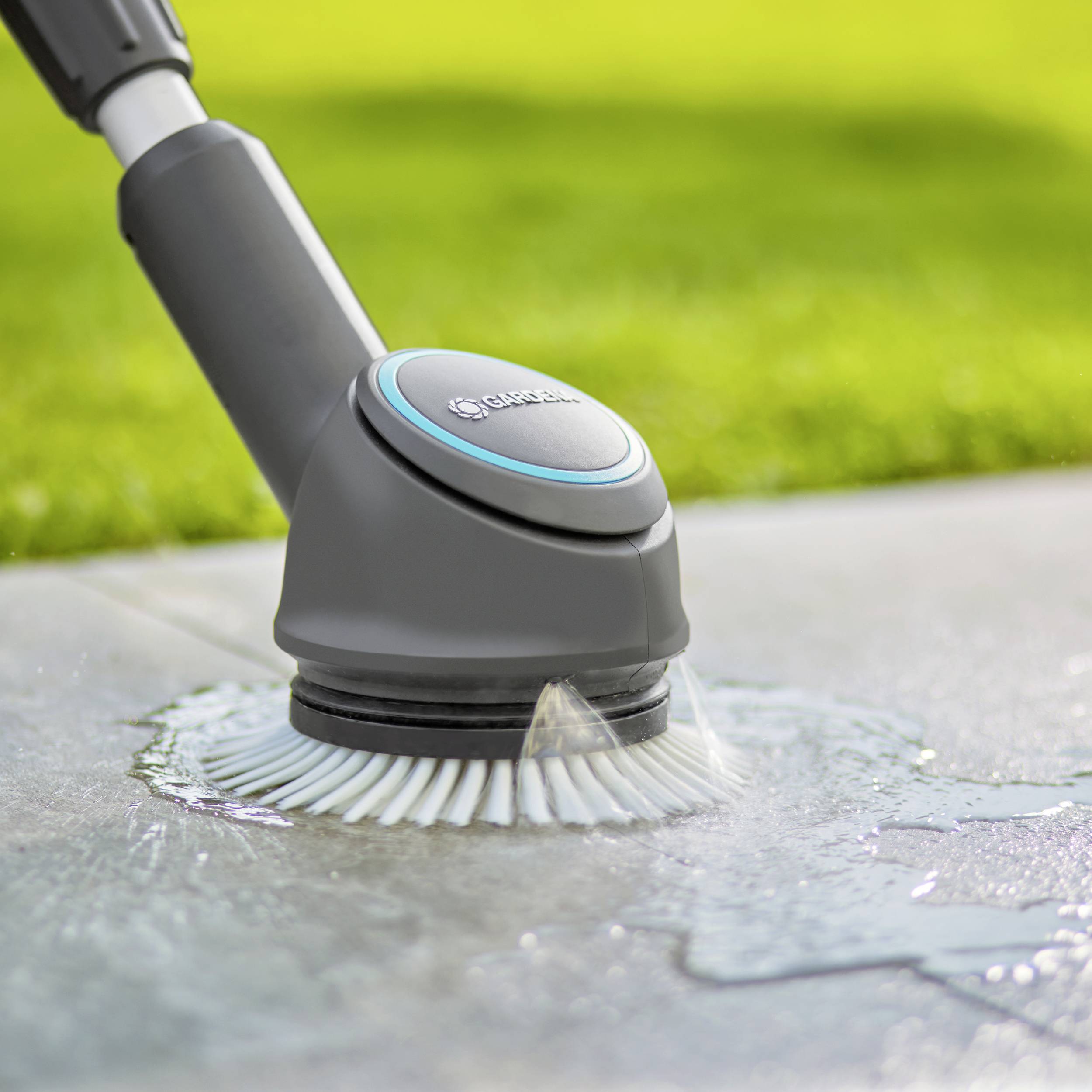 An electric cleaning brush cleans a smooth, wet surface outdoors with water. Grass is visible in the background.
