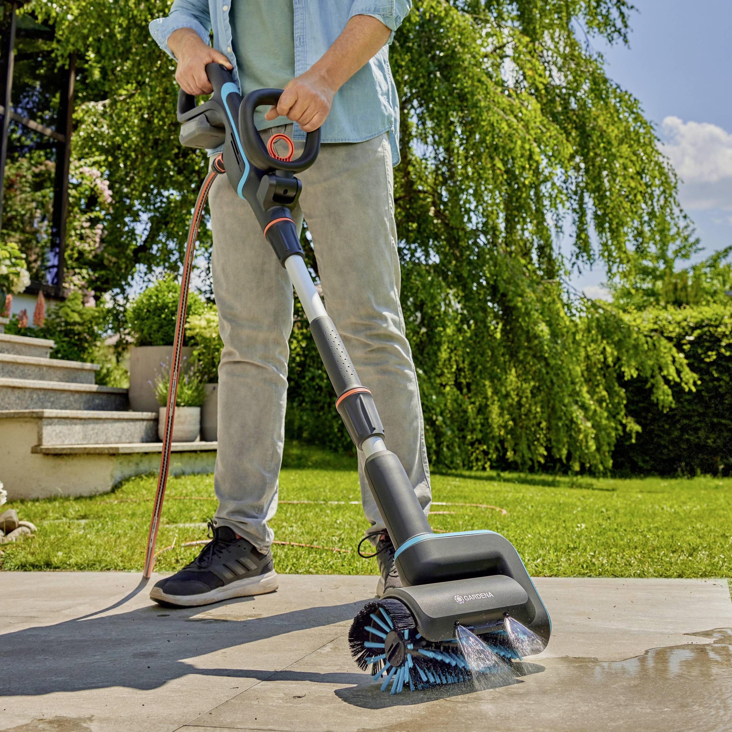 A person is cleaning a paved patio with an electric cleaning device that sprays water. Garden in the background.