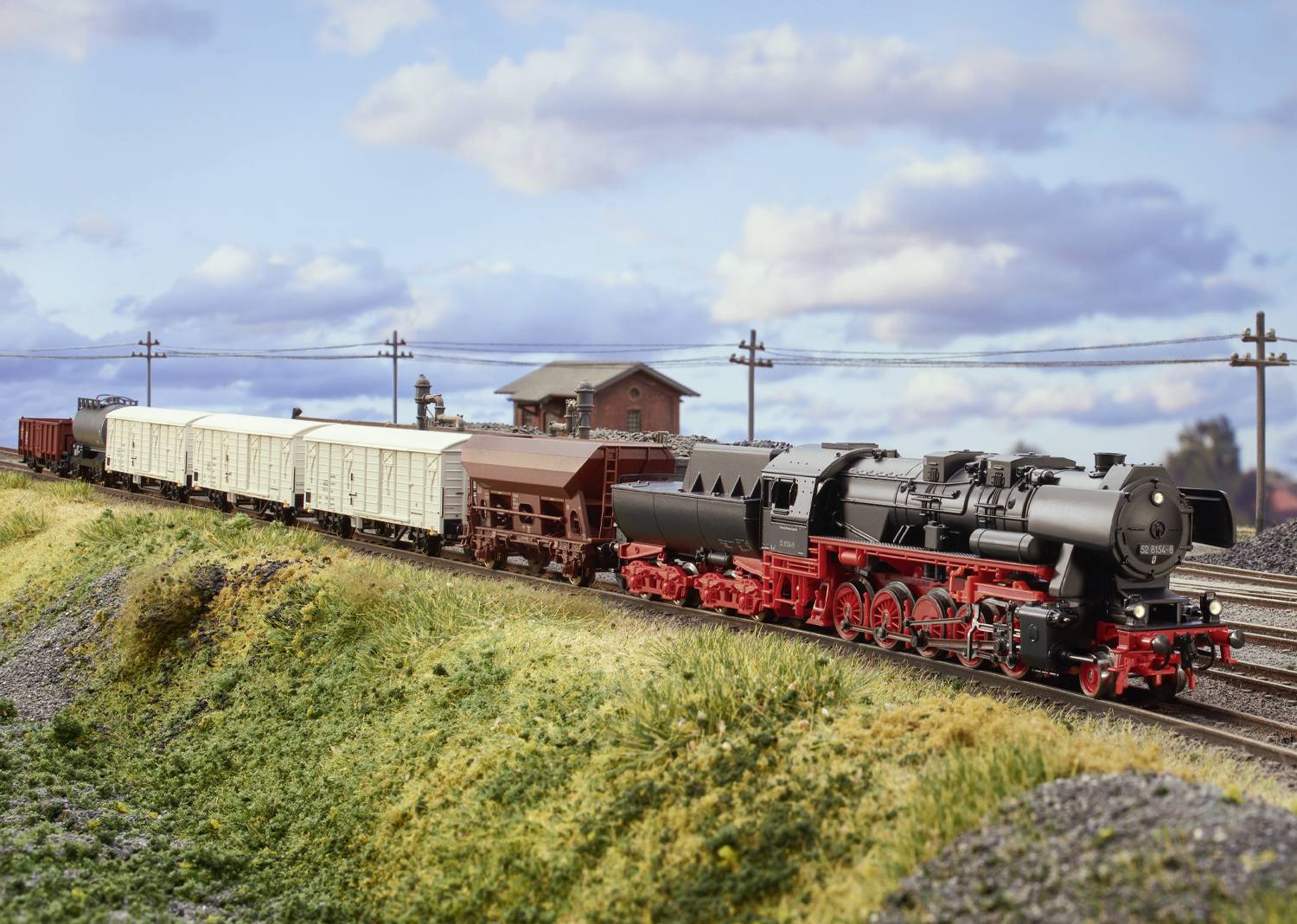 A steam locomotive is pulling goods wagons on a model railway through a landscape with meadows and a building in the background.