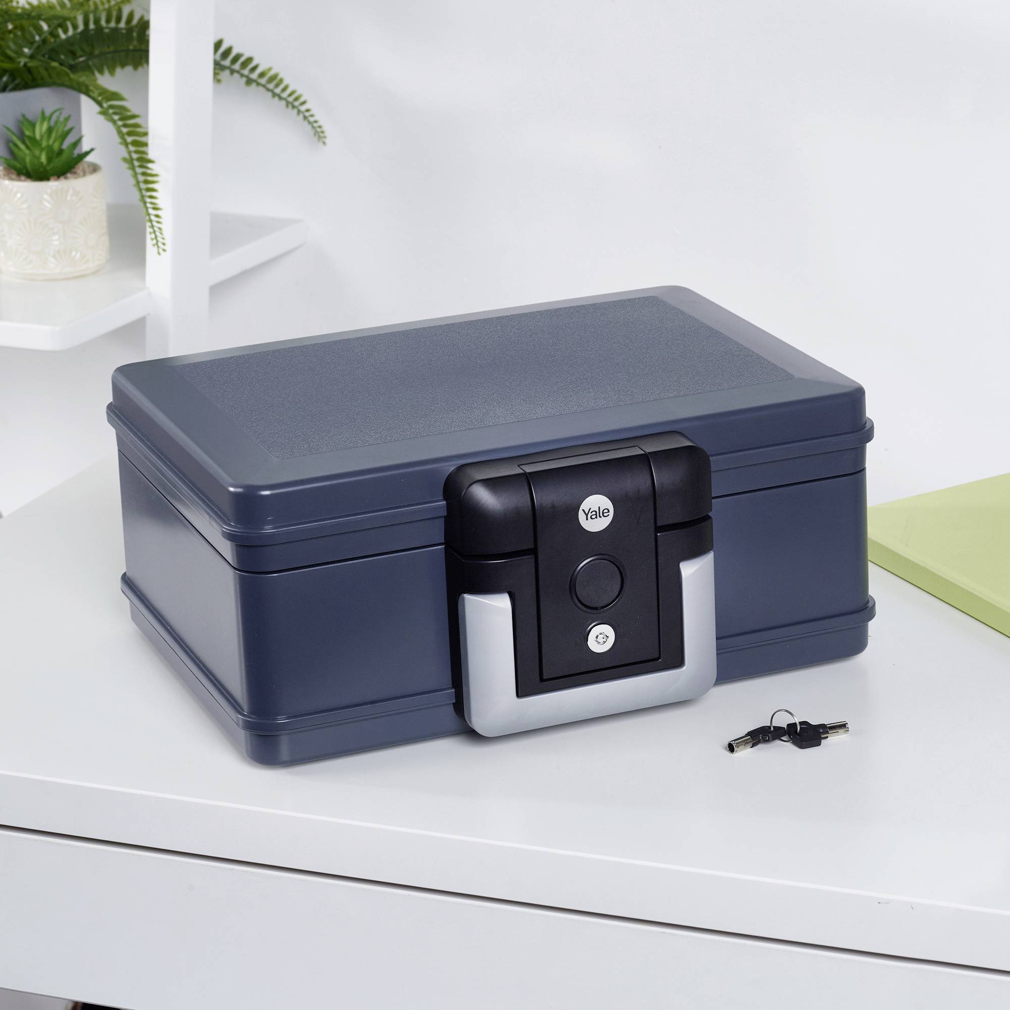 A blue-grey safe on a desk with plants in the background. Keys are lying beside it.