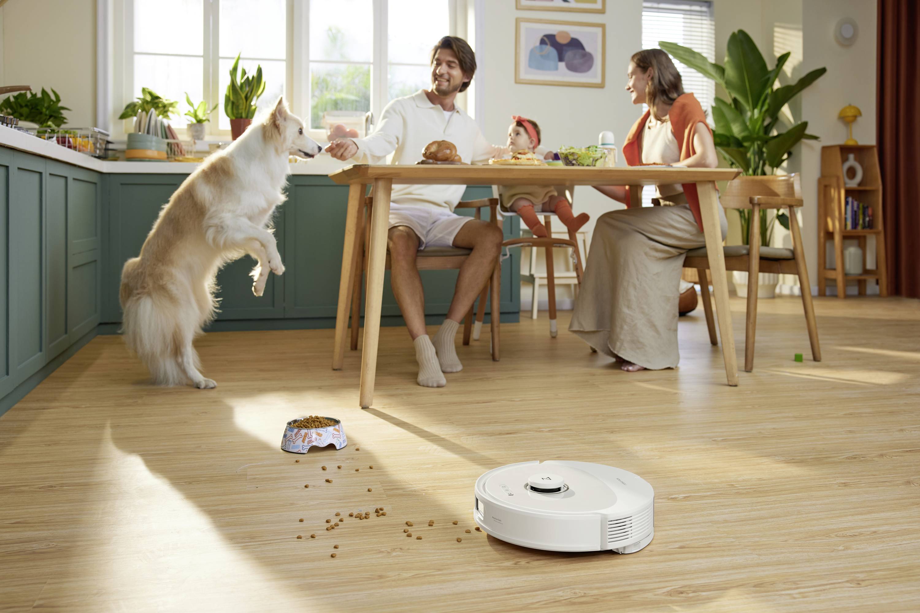 A dog jumps up enthusiastically while a couple sits at the table chatting. A baby in a high chair is laughing. A robotic vacuum cleaner is cleaning.