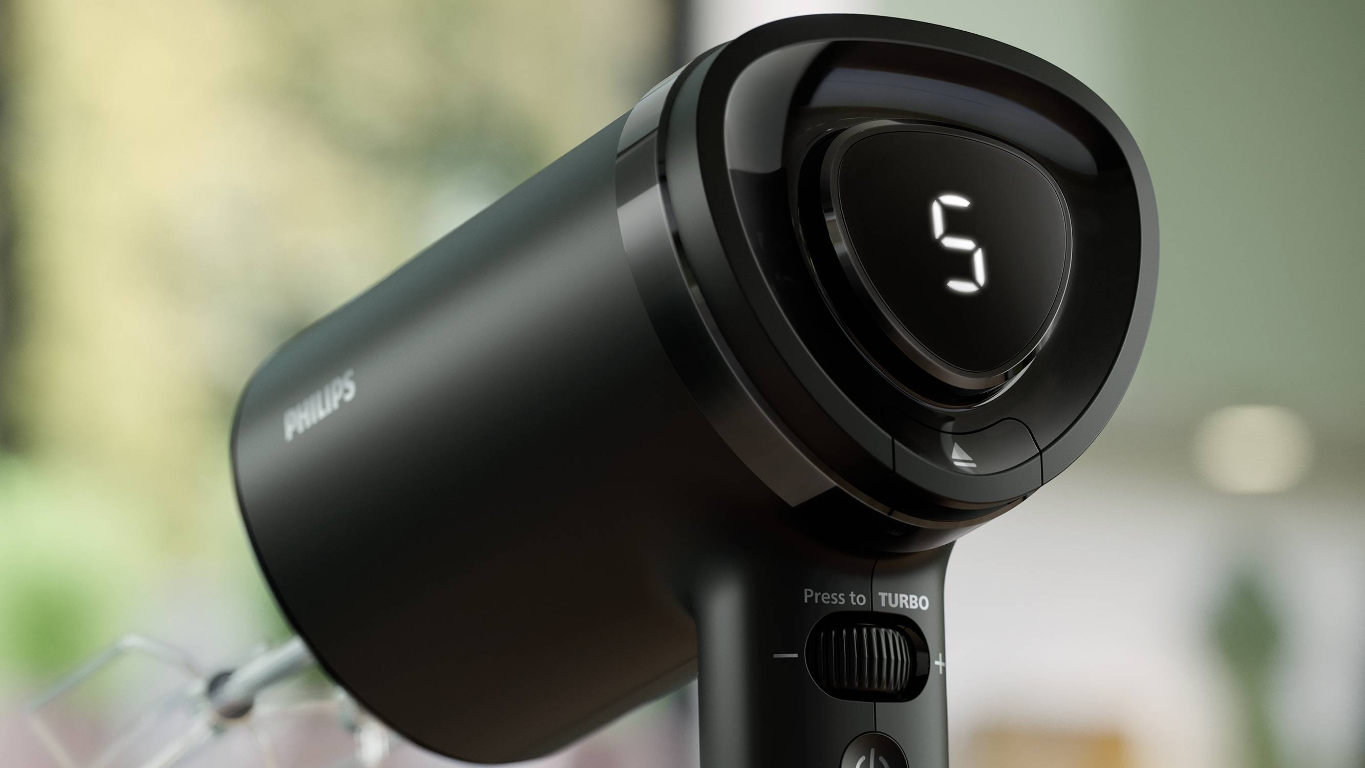 Black hairdryer with digital display '5'. It is standing on a table, surrounded by a blurred green environment in the background.