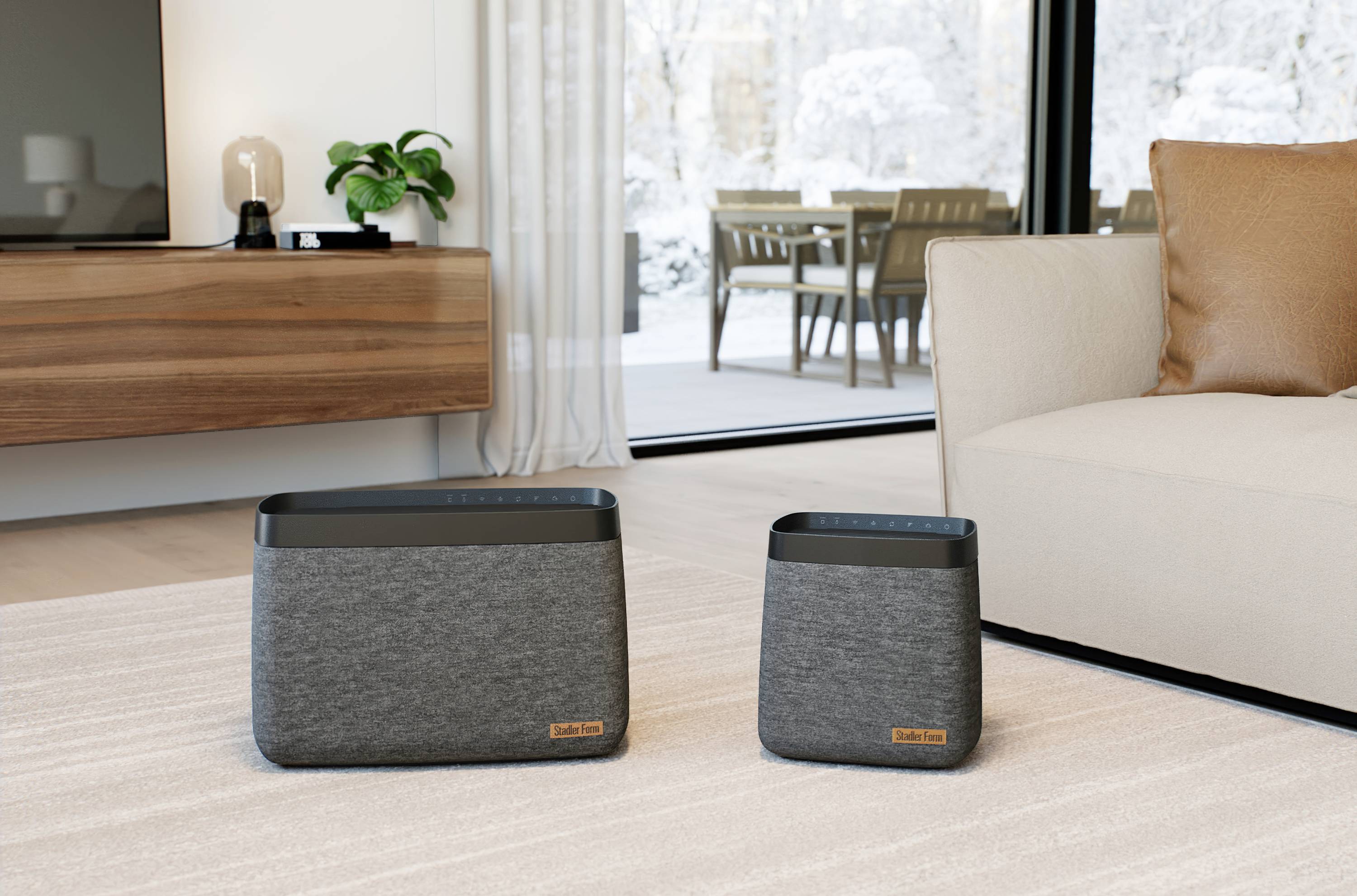 Two grey speakers of different sizes stand on a carpet in a modern living room with a view of a terrace in winter.