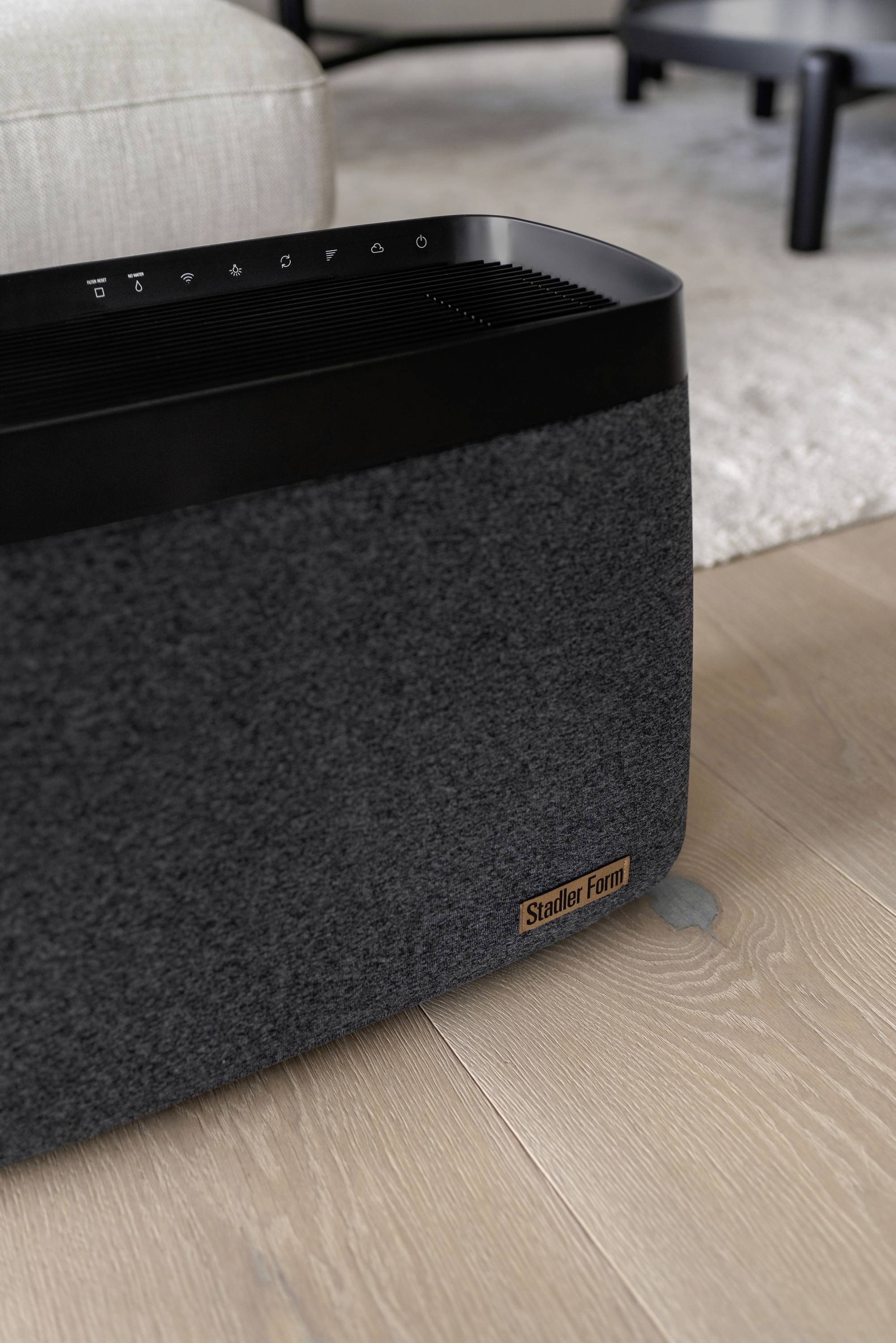 A grey home speaker stands on a wooden floor. The speaker has a rectangular shape and is dark with a fabric cover.