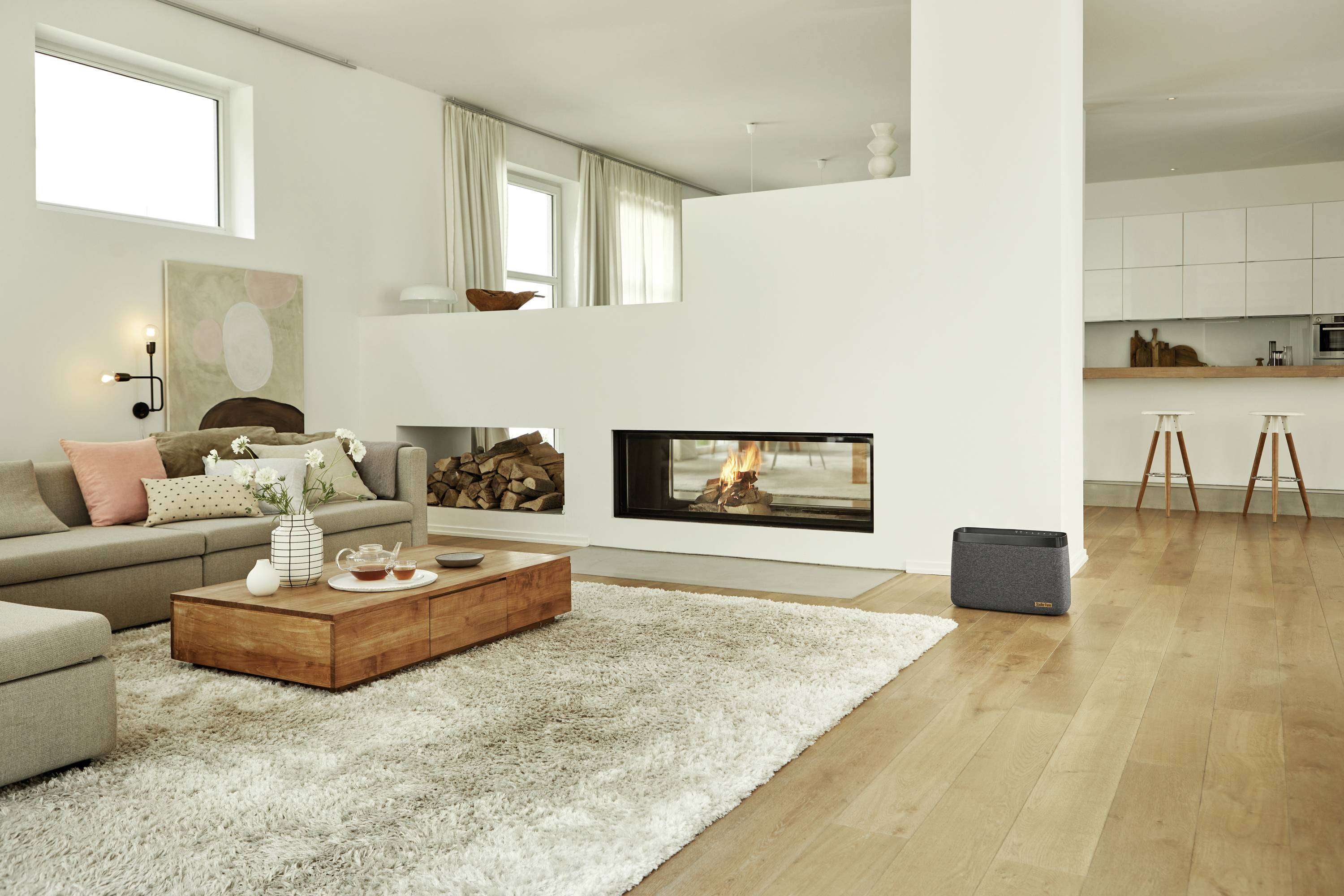 Cosy living room with a fireplace, light wooden flooring and modern furnishings. Open-plan kitchen in the background.