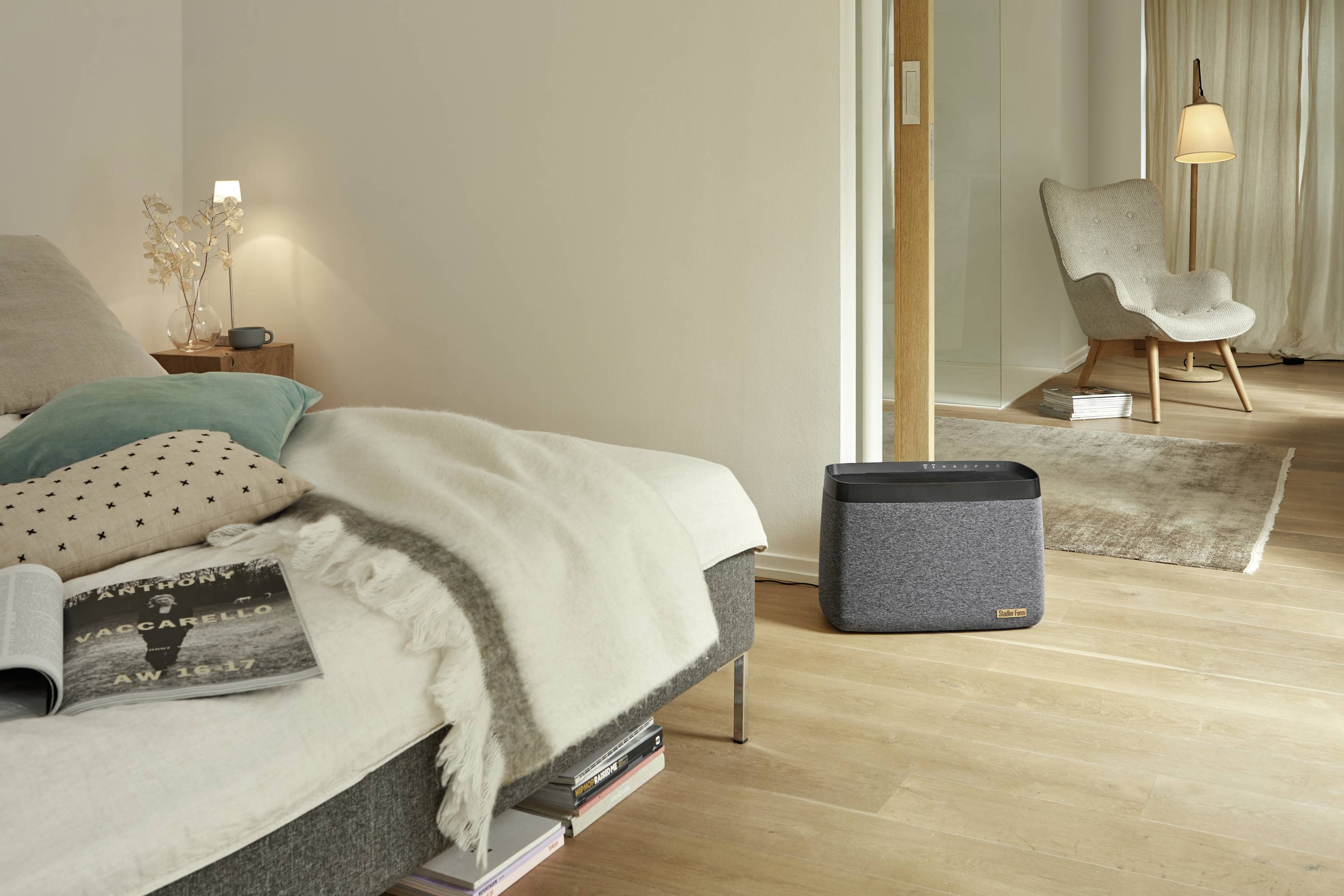 A modern furnished bedroom with a grey speaker on the floor, a bed with pillows and blankets on the left, and an armchair on the right.