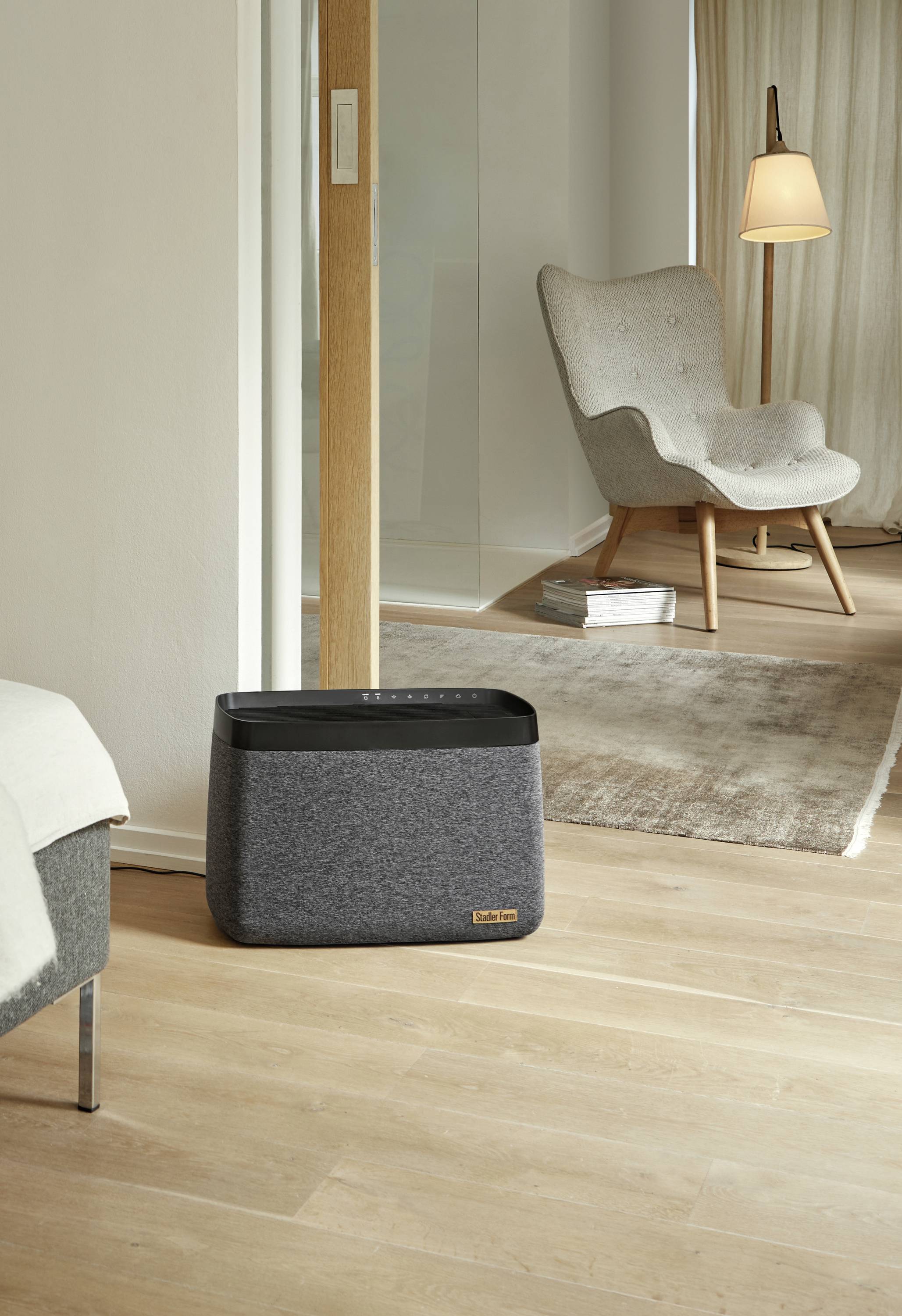 A grey, cuboid air purifier stands in a modern living room in front of an armchair and a lamp on a light wooden floor.