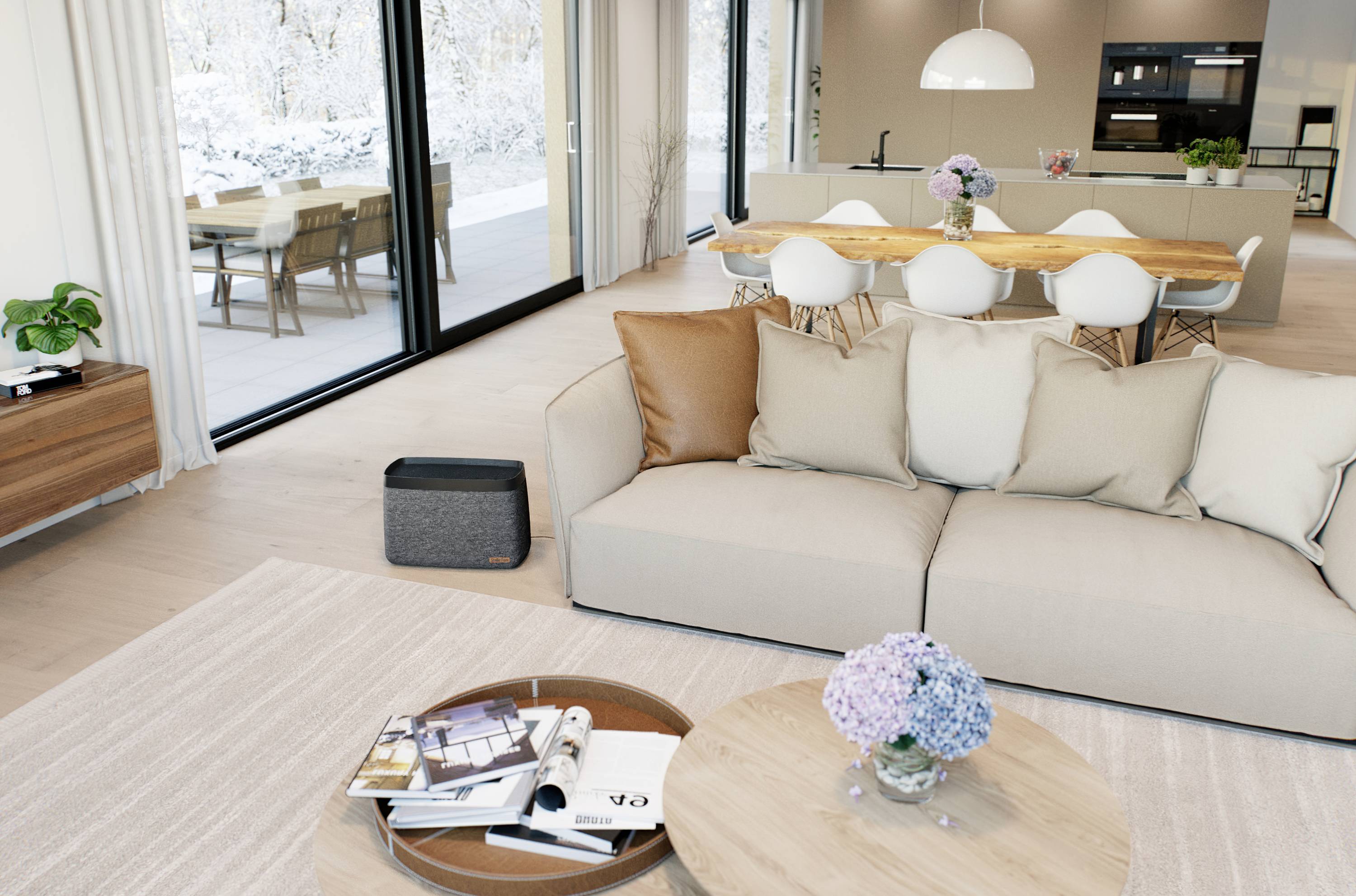 Modern living room with a beige sofa, coffee table with flowers, view of kitchen and dining area with large windows overlooking the snow-covered exterior.